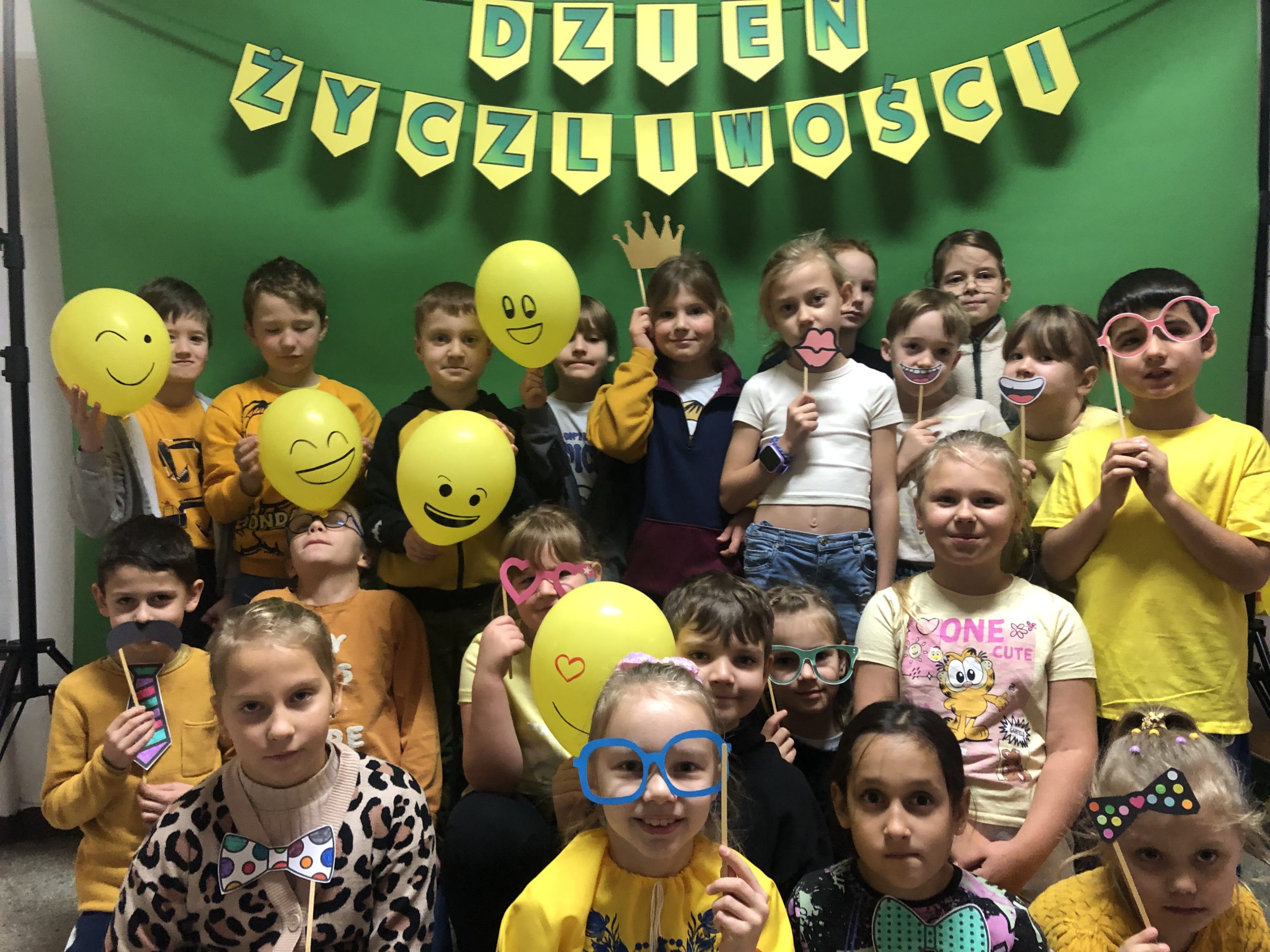 Grupa dzieci ubranych na żółto pozuje z papierowymi emotikonami, balonami i akcesoriami do zdjęć z okazji „Dnia Życzliwości”, stojąc przed zielonym tłem z dekoracyjnym napisem DZIEŃ ŻYCZLIWOŚCI