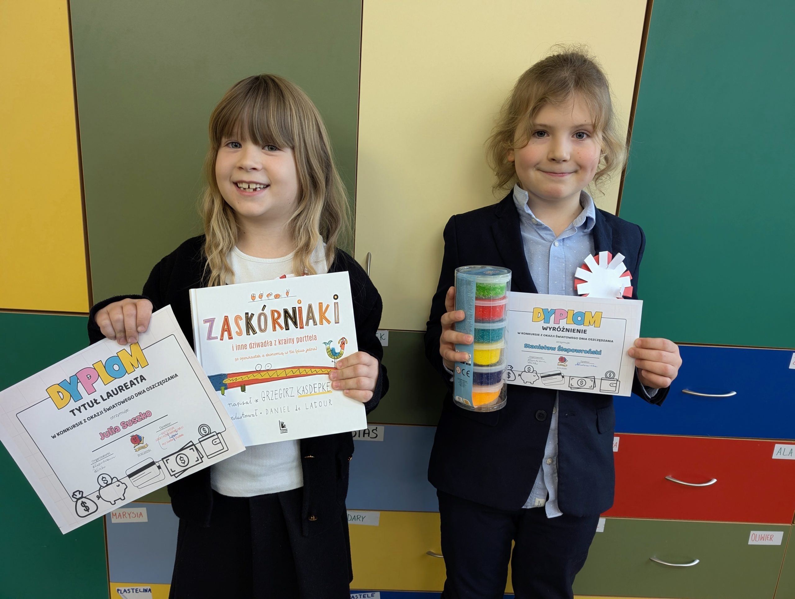 Od lewej uśmiechnięta dziewczynka i chłopiec z klasy 2d. Trzymają w rękach dyplomy, dziewczynka dodatkowo trzyma książkę, a chłopiec masę plastyczną w pudełkach. W tle szkolne szafki.