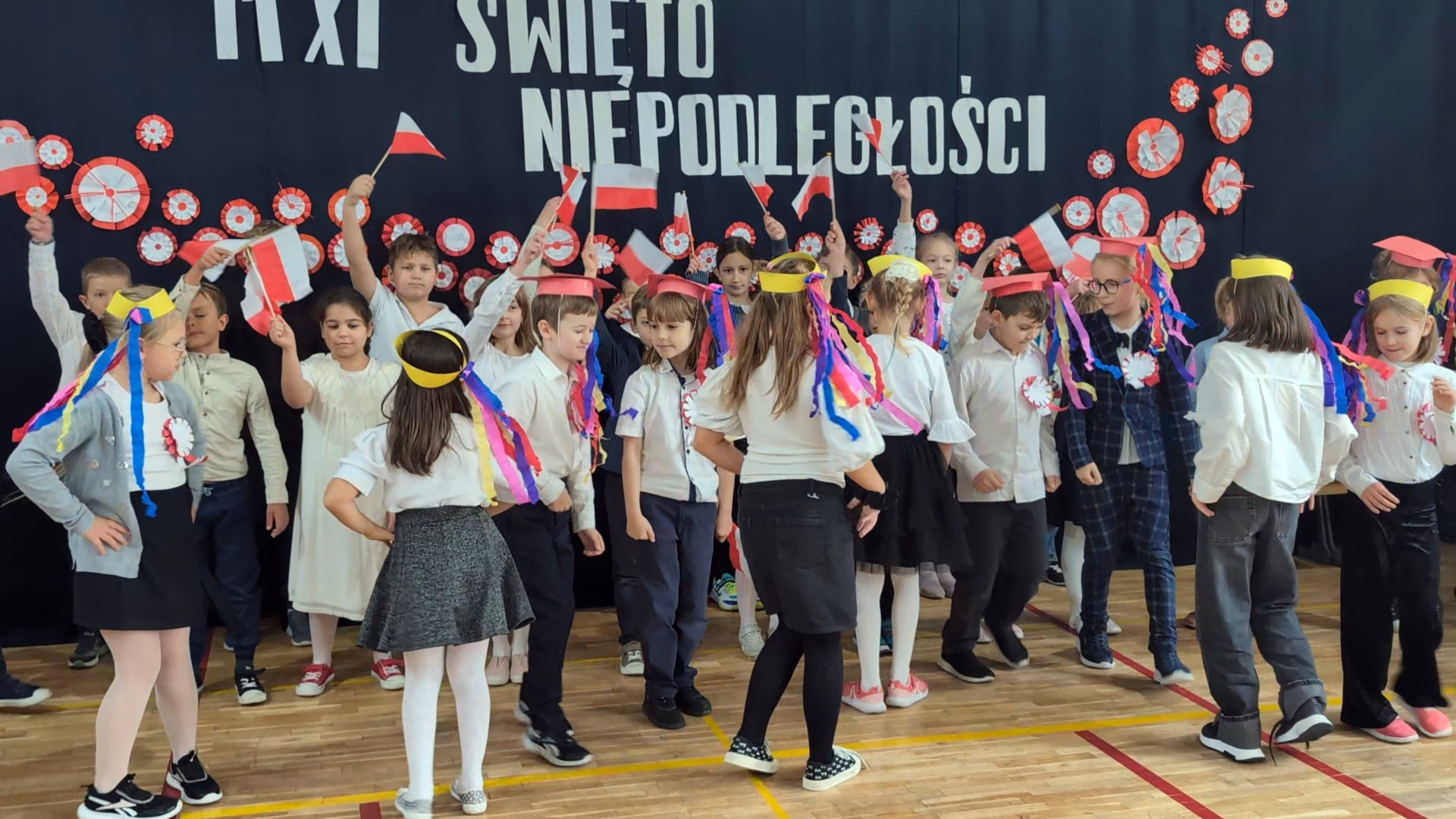 Dzieci w szkolnej sali gimnastycznej świętują Dzień Niepodległości. Na tle dekoracji z napisem „11 XI ŚWIĘTO NIEPODLEGŁOŚCI” trzymają biało-czerwone flagi i noszą papierowe czapki z kolorowymi wstążkami. Niektóre dzieci stoją w szeregu, inne tańczą lub śpiewają.