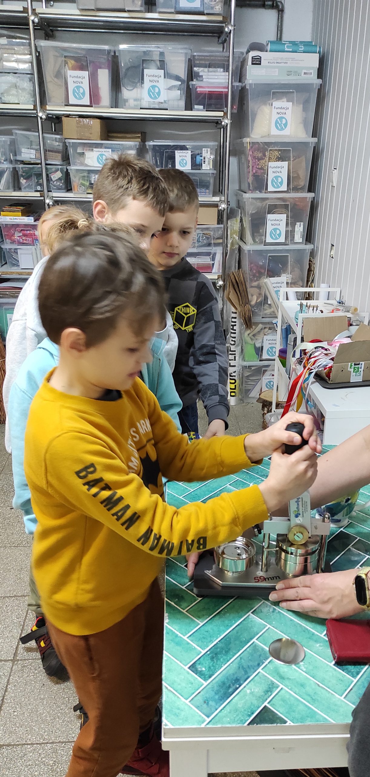 Na pierwszym planie chłopiec obsługujący maszynę do przypinek. W tle dłonie dorosłej osoby oraz inne dzieci i elementy wysposażenia sali.