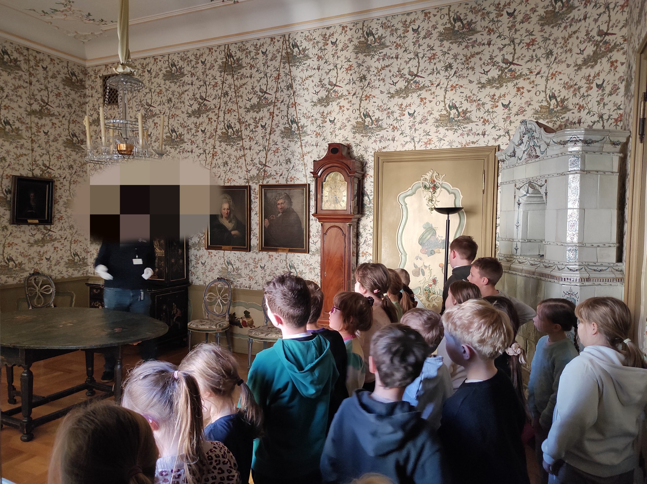 17 uczniów stoi w elegancko urządzonym pomieszczeniu. Przodem do dzieci stoi  mężczyzna. Na ścianach tapeta w liście i ptaki oraz 3 obrazy. W pieszczeniu znajduje się stół, piec kaflowy i zegar stojący.