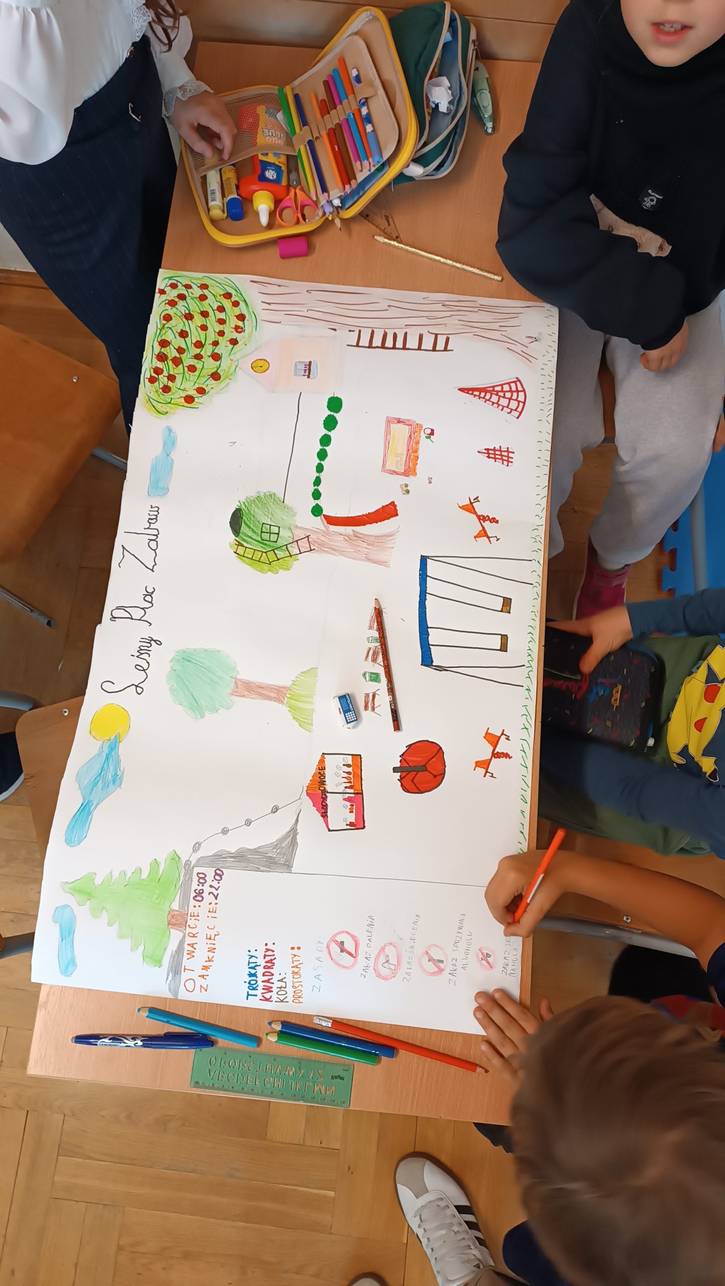 Dzieci pracują nad dużym plakatem przedstawiającym projekt placu zabaw. Na stole leżą kredki, flamastry i przybory plastyczne, a na kartce widać kolorowe rysunki drzew, huśtawek i boiska.