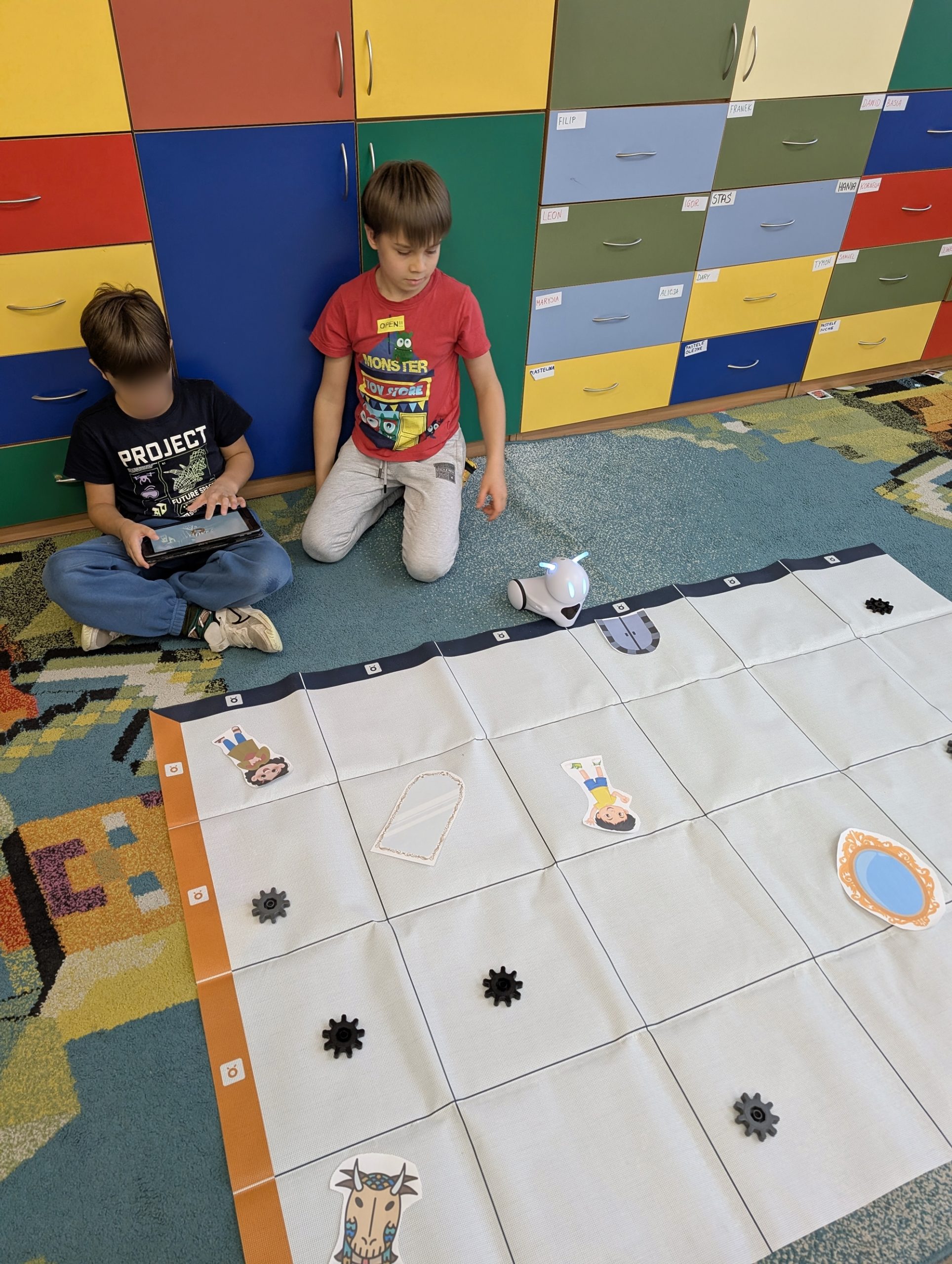 Dwóch chłopców siedzi na dywanie w klasie przy dużej macie do programowania. Na macie leżą ilustracje postaci oraz przeszkody, a obok nich stoi mały robot sterowany tabletem.