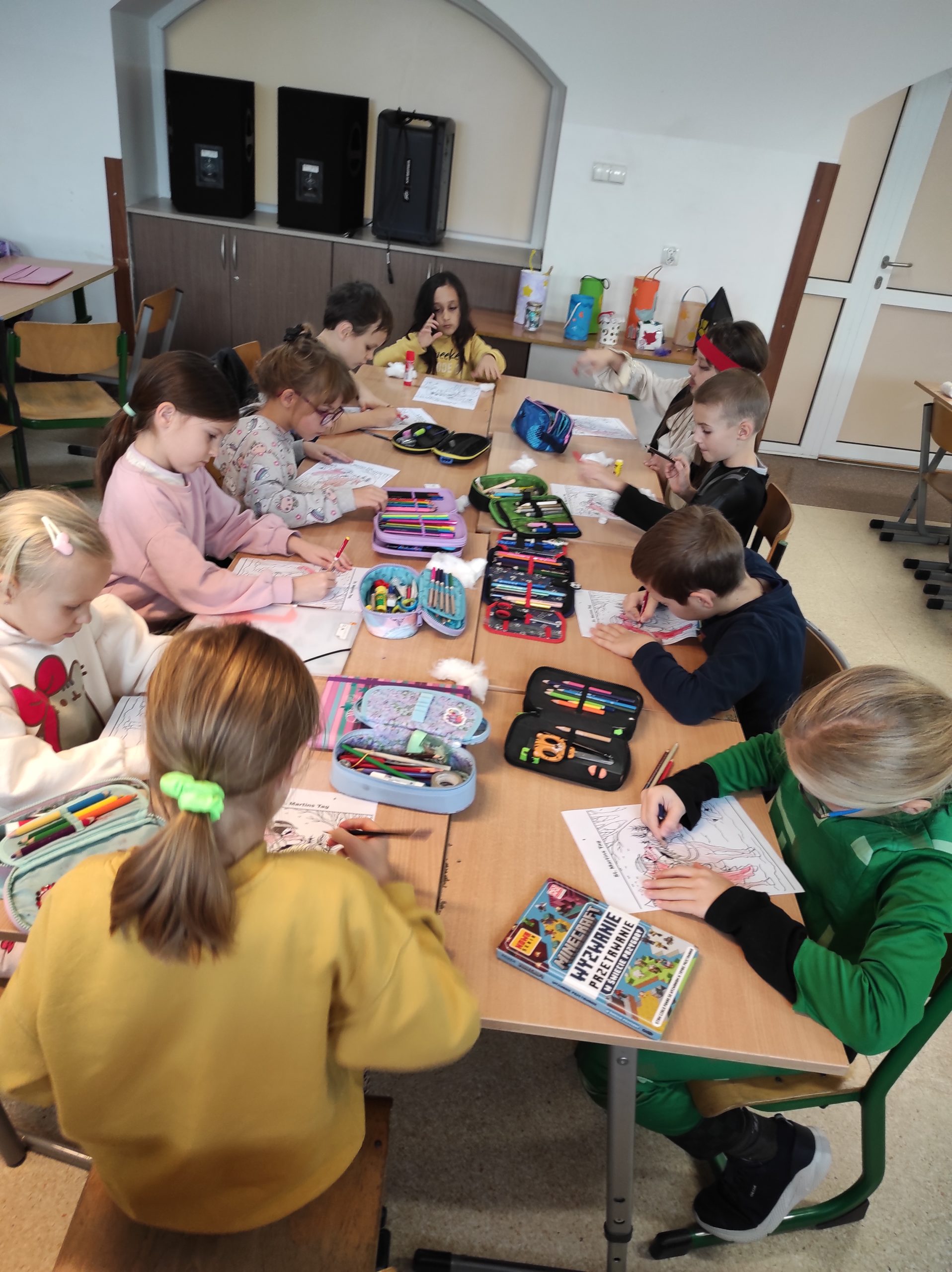 Uczniowie siedzą przy stolikach, na których leżą piórniki, kleje, wata, kartki. W tle elementy sali lekcyjnej.