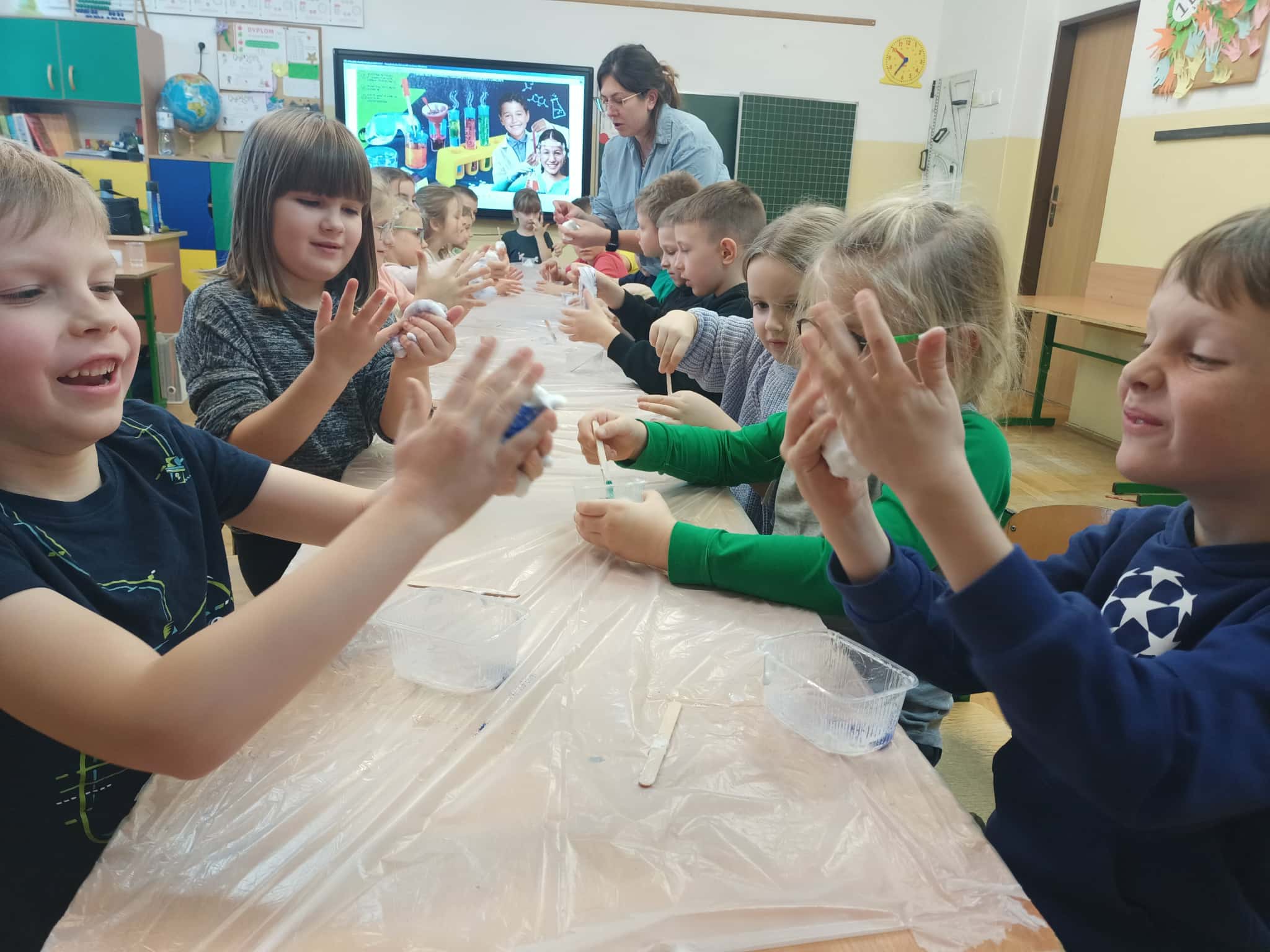 Grupa uczestników zajęć przy długim stole ugniata i roluje slime w dłoniach, a nauczycielka pomaga w tle; na tablicy multimedialnej wyświetlona jest prezentacja o doświadczeniach.