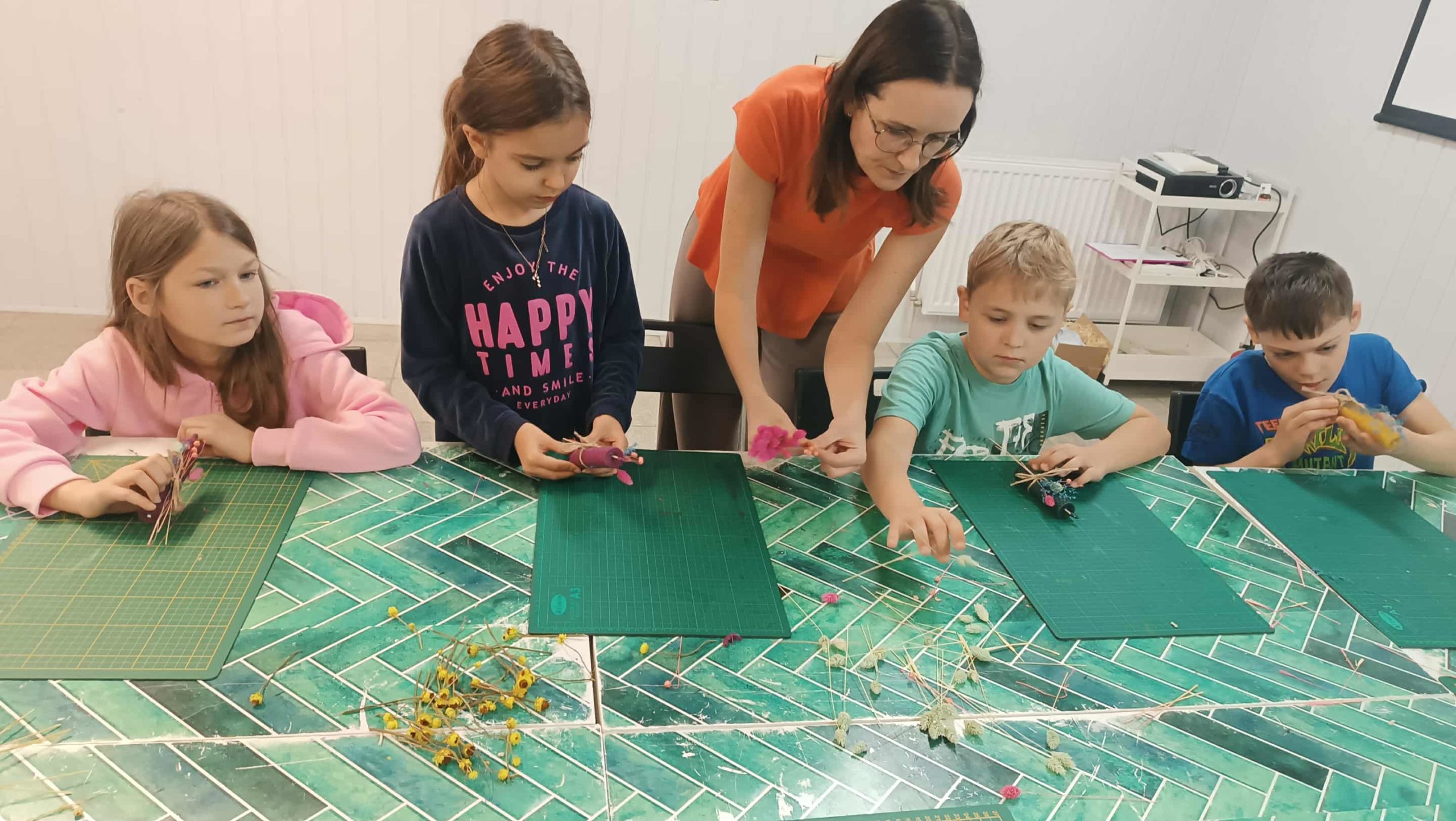Dzieci siedzą przy stole i ozdabiają świece suszonymi roślinami oraz materiałów dekoracyjnych. Obok nich stoi instruktorka, która pomaga jednej z dziewczynek przy wykonaniu świecy. Na stole leżą maty do cięcia i rozłożone suszone kwiaty.