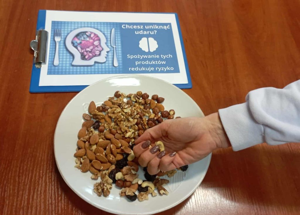 Na stole leży grafika dotycząca Udaru Mózgu. Na jej tle dłoń sięgająca po orzechy z talerza
