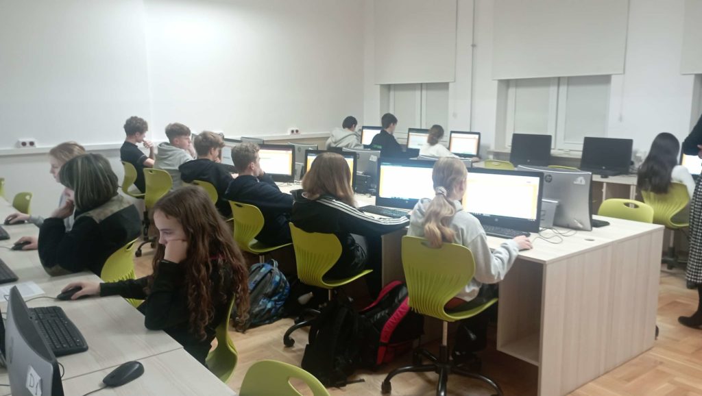 W sali komputerowej na tle okien przy komputerach siedzi 13 uczniów klas starszych. Na pierwszym planie rząd monitorów stojących na biurkach.