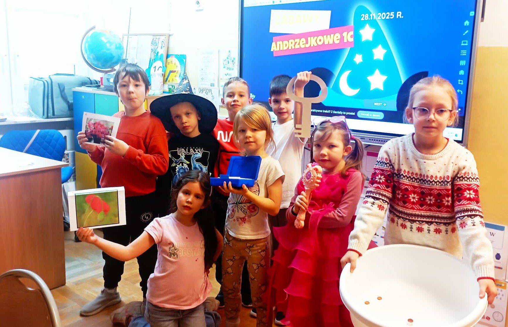 8 uczniów klasy 1c pozuje do zdjęcia w sali na tle tablicy multimedialnej z napisem ZABAWY ANDRZEJKOWE 1c. W rękach trzymją akcesoria do zabawy we wróżby.