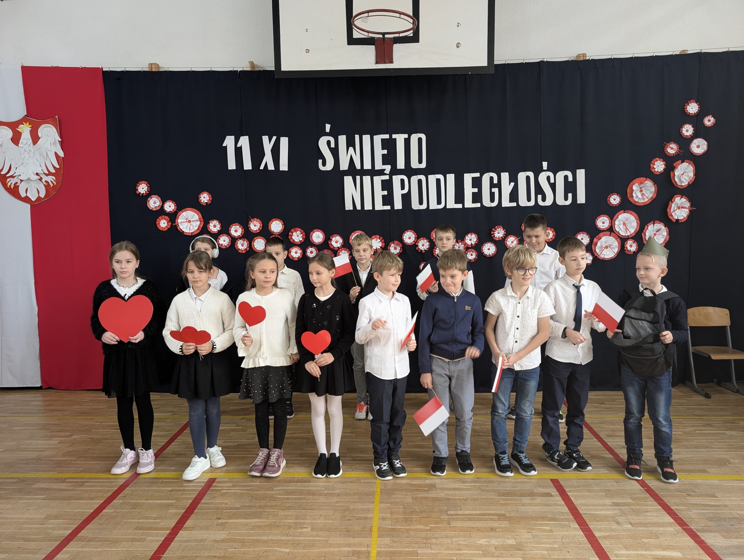 Uczniowie z klasy 3a stoją na sali gimnastycznej przed dekoracją z napisem „11 XI Święto Niepodległości”. Dzieci ubrane są odświętnie, a w dłoniach trzymają flagi, serca, a chłopiec po prawej  plecak, bo śpiewają pieśń "Serce w  plecaku".
