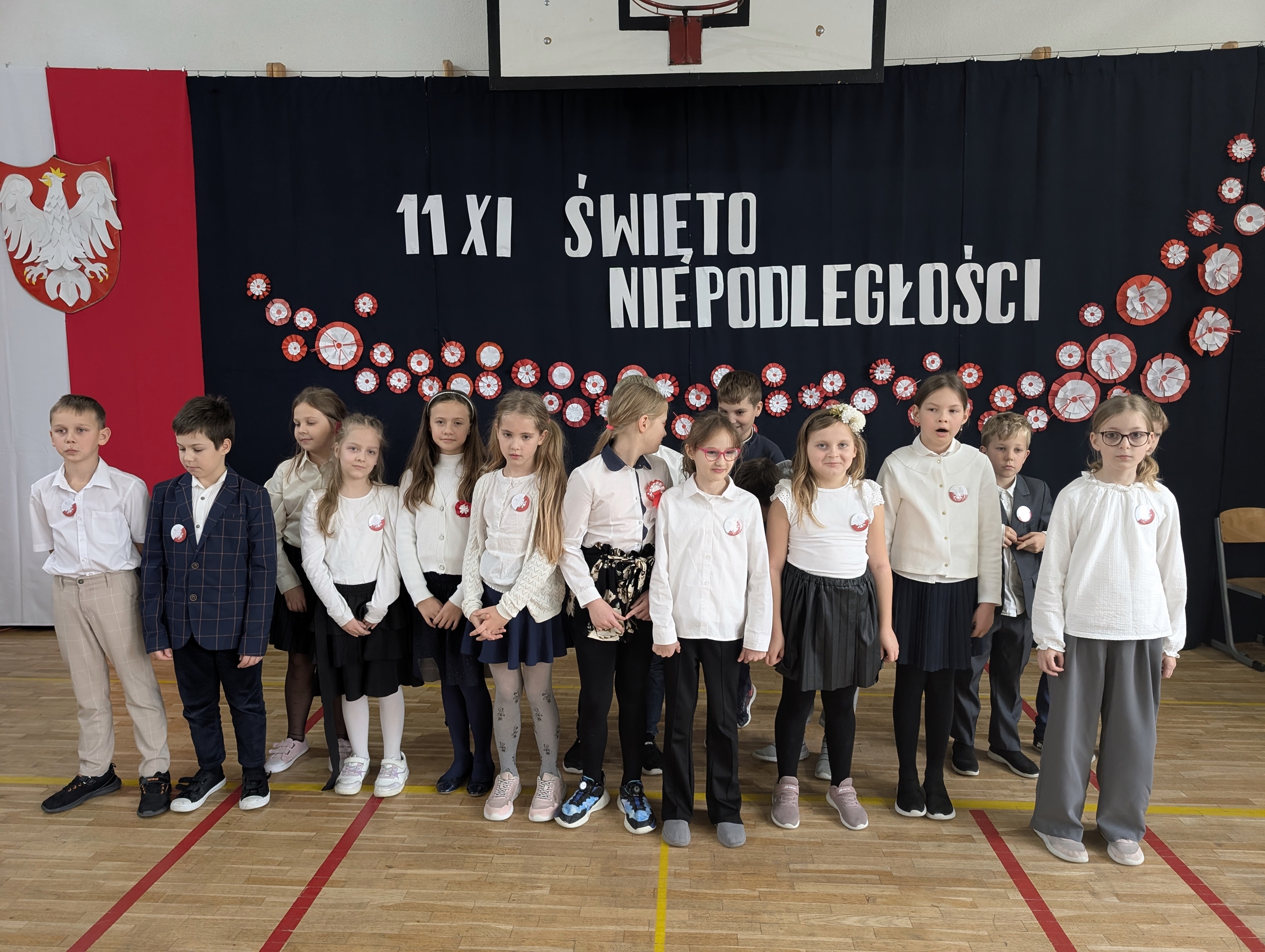 Uczniowie z klasy 2b stoją na sali gimnastycznej przed dekoracją z napisem „11 XI Święto Niepodległości”. Dzieci ubrane są odświętnie, mają przyczepione do ubrań kokardy narodowe.