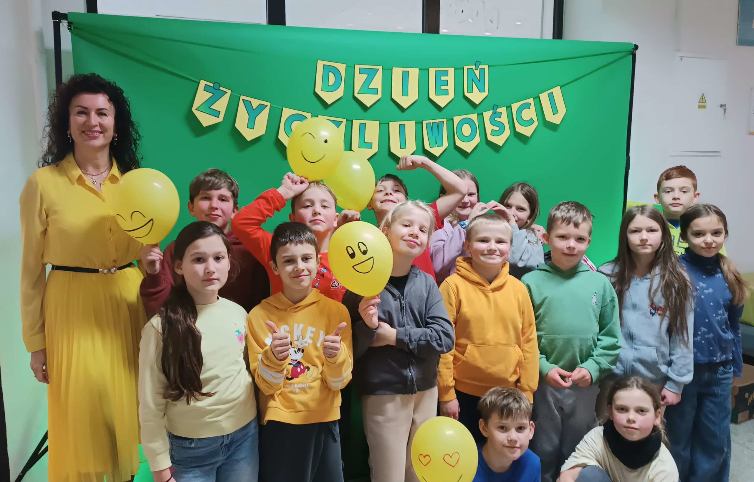 Grupa dzieci ubranych na żółto pozuje z papierowymi emotikonami, balonami i akcesoriami do zdjęć z okazji „Dnia Życzliwości”, stojąc przed zielonym tłem z dekoracyjnym napisem DZIEŃ ŻYCZLIWOŚCI