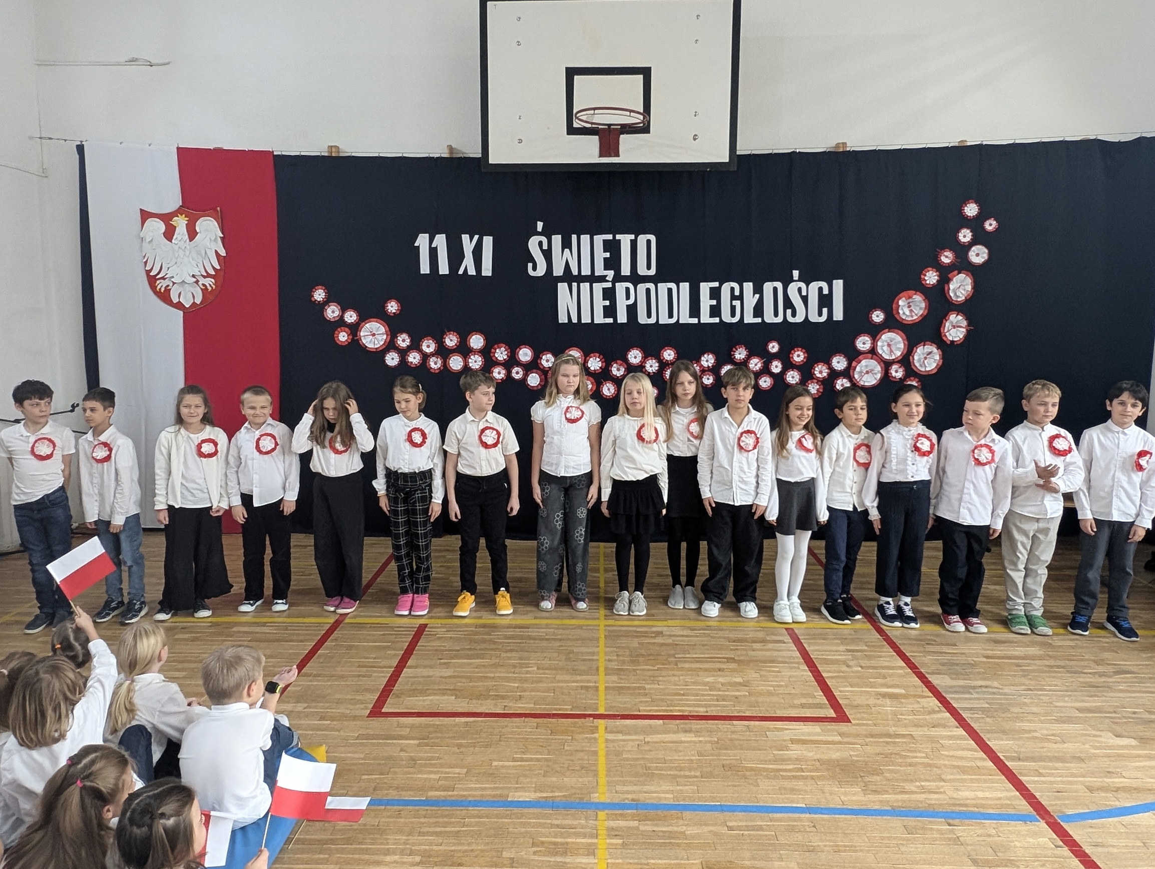 Uczniowie z klasy 3c stoją w szeregu na sali gimnastycznej przed dekoracją z napisem „11 XI Święto Niepodległości”. Dzieci ubrane są odświętnie, mają przyczepione do ubrań kokardy narodowe.