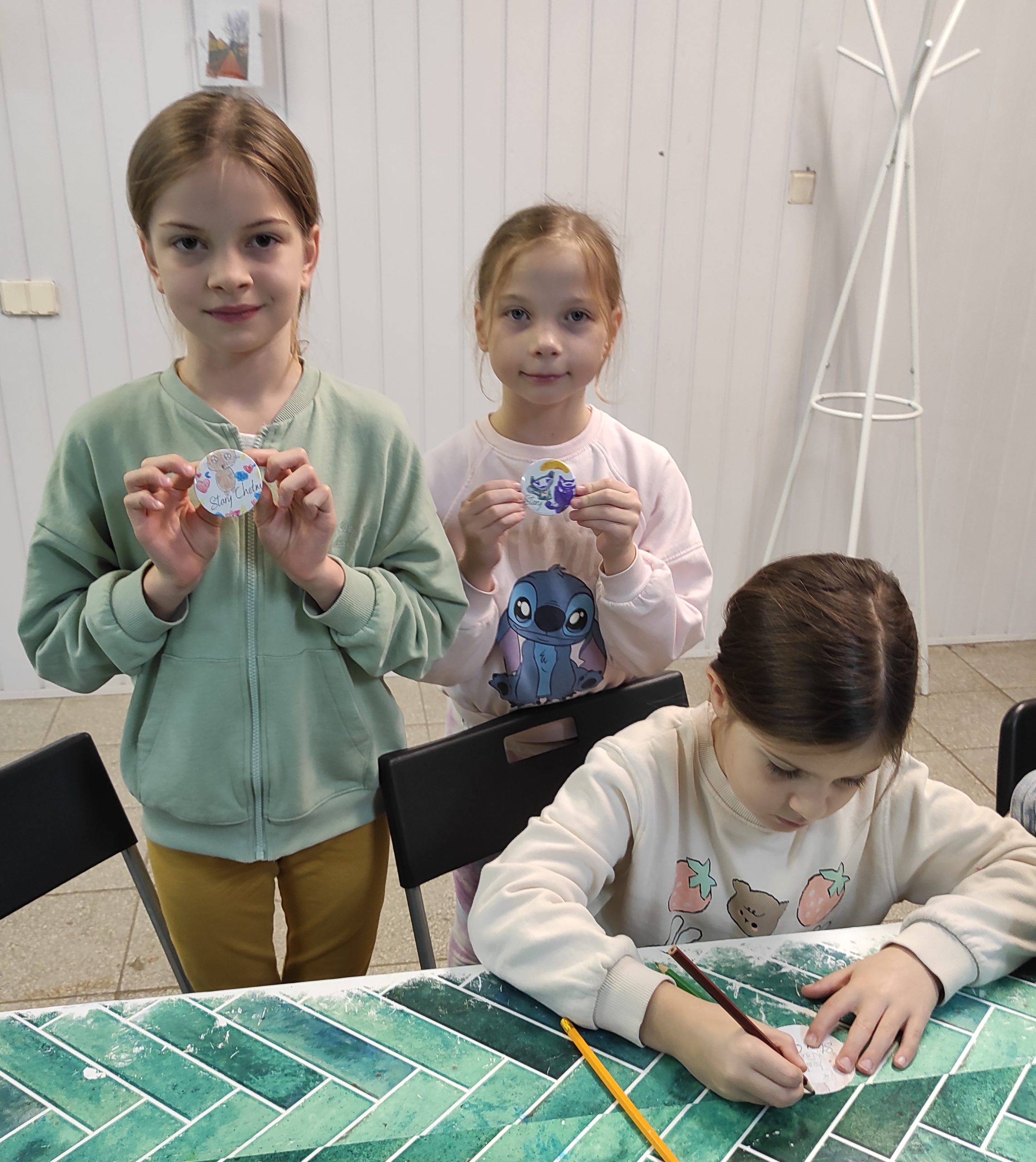 Na pierwszym planie trzy dziewczynki: jedna siedzi przy stole i ma w prawej dłoni kredkę, a lewą przytrzymuje małą karteczkę, dwie pozostałe dziewczynki stoją przy niej i pokazują przypinki.