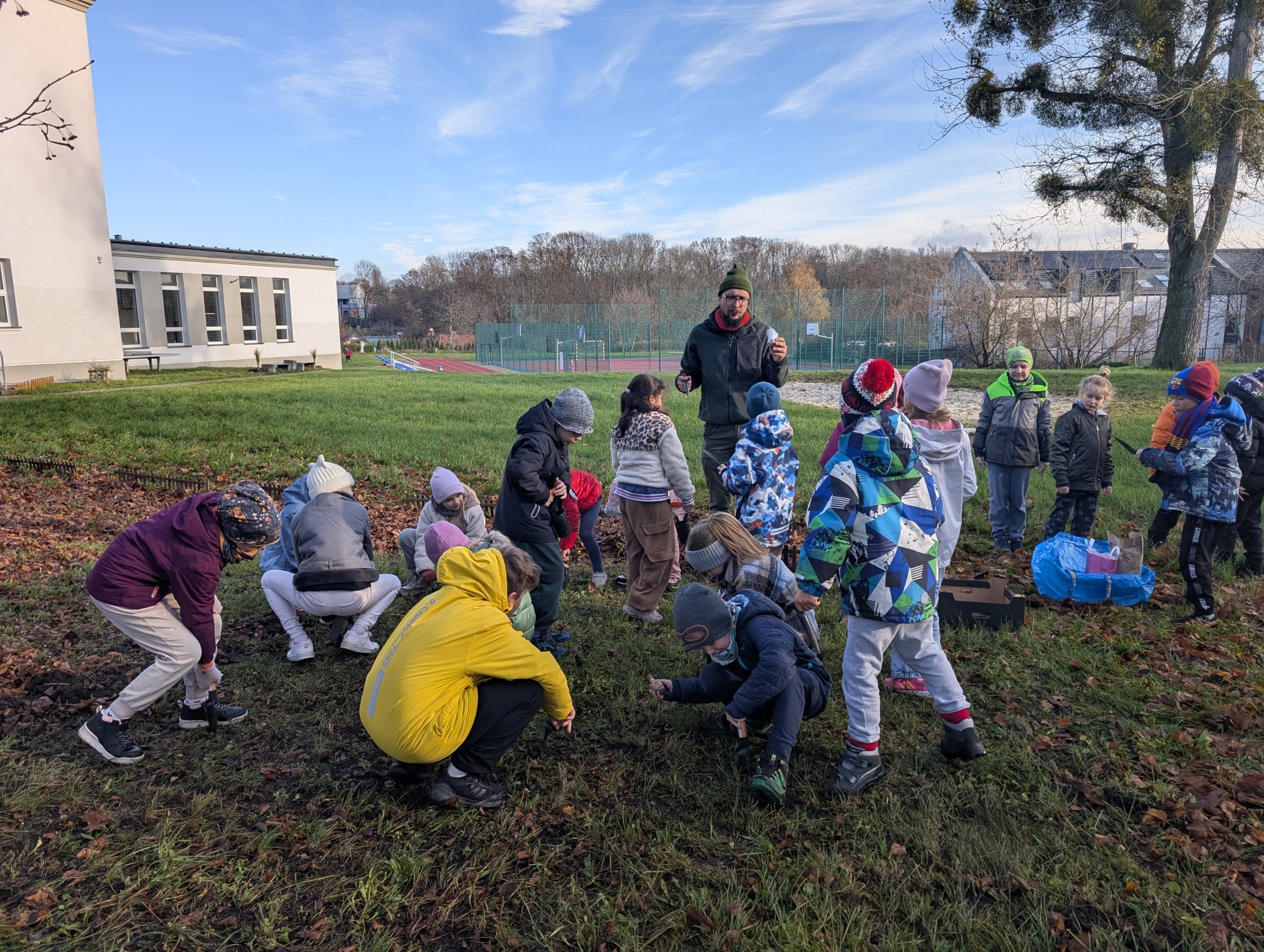 Grupa dzieci stoi w półkolu na trawniku, obserwując osobę dorosłą klęczącą przy skrzynce z narzędziami ogrodniczymi. W tle widać boisko, drzewa oraz sprzęty plenerowe.