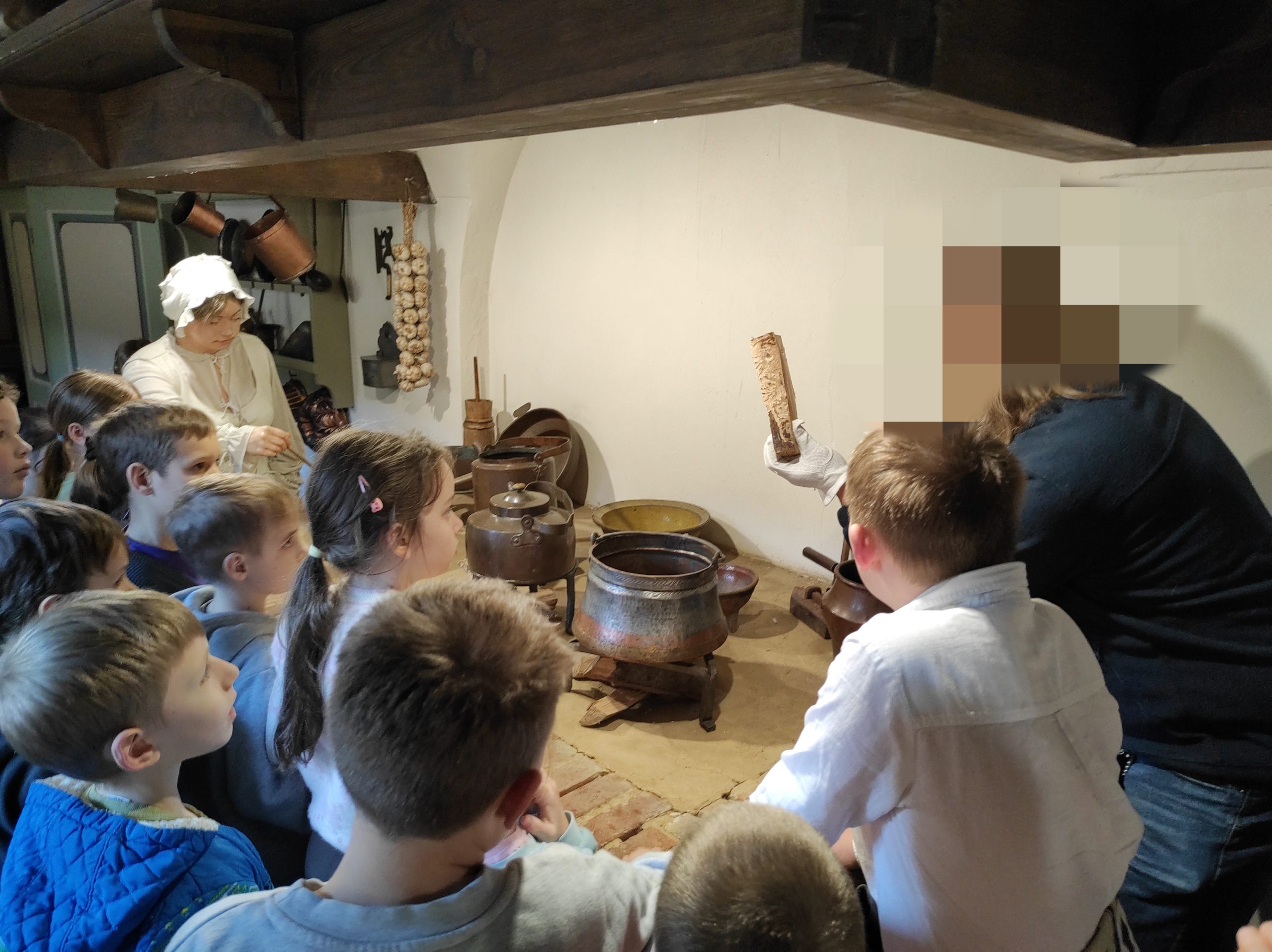 10 uczniów, mężczyzna i manekin ubrany jak gosposia stoją w pomieszczeniu. Wszyscy patrzą w stronę naczyń leżących przed nimi. Mężyczna trzyma w dłoni odłupany kawałek drewna. 