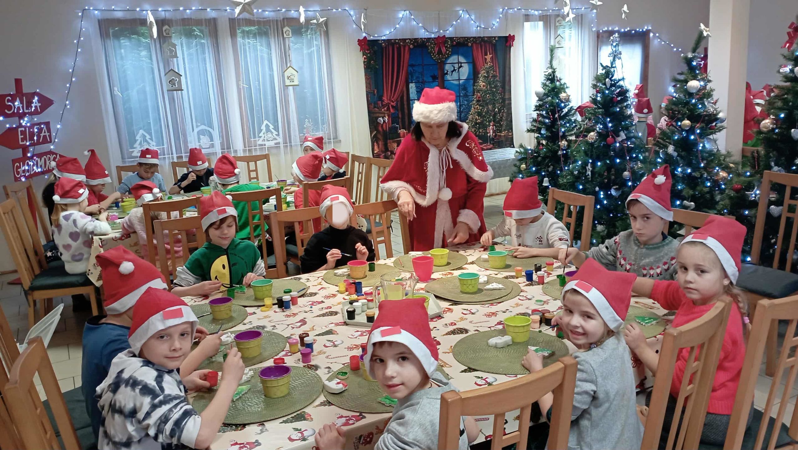 Grupa dzieci w czerwonych czapkach Mikołaja siedzi przy dużych, stołach i wykonuje świąteczne prace plastyczne. W sali znajdują się ozdobione choinki z lampkami, a w tle stoi osoba przebrana w strój świąteczny, pomagająca dzieciom.
