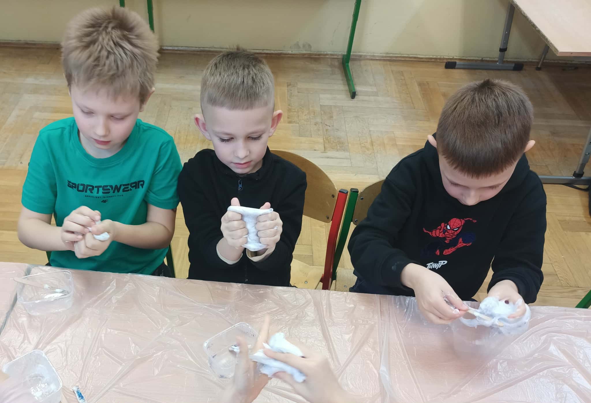 Trzech chłopców przy stoliku ugniata i rozciąga białą masę slime; na blacie widać plastikowe pojemniki i patyczki do mieszania.