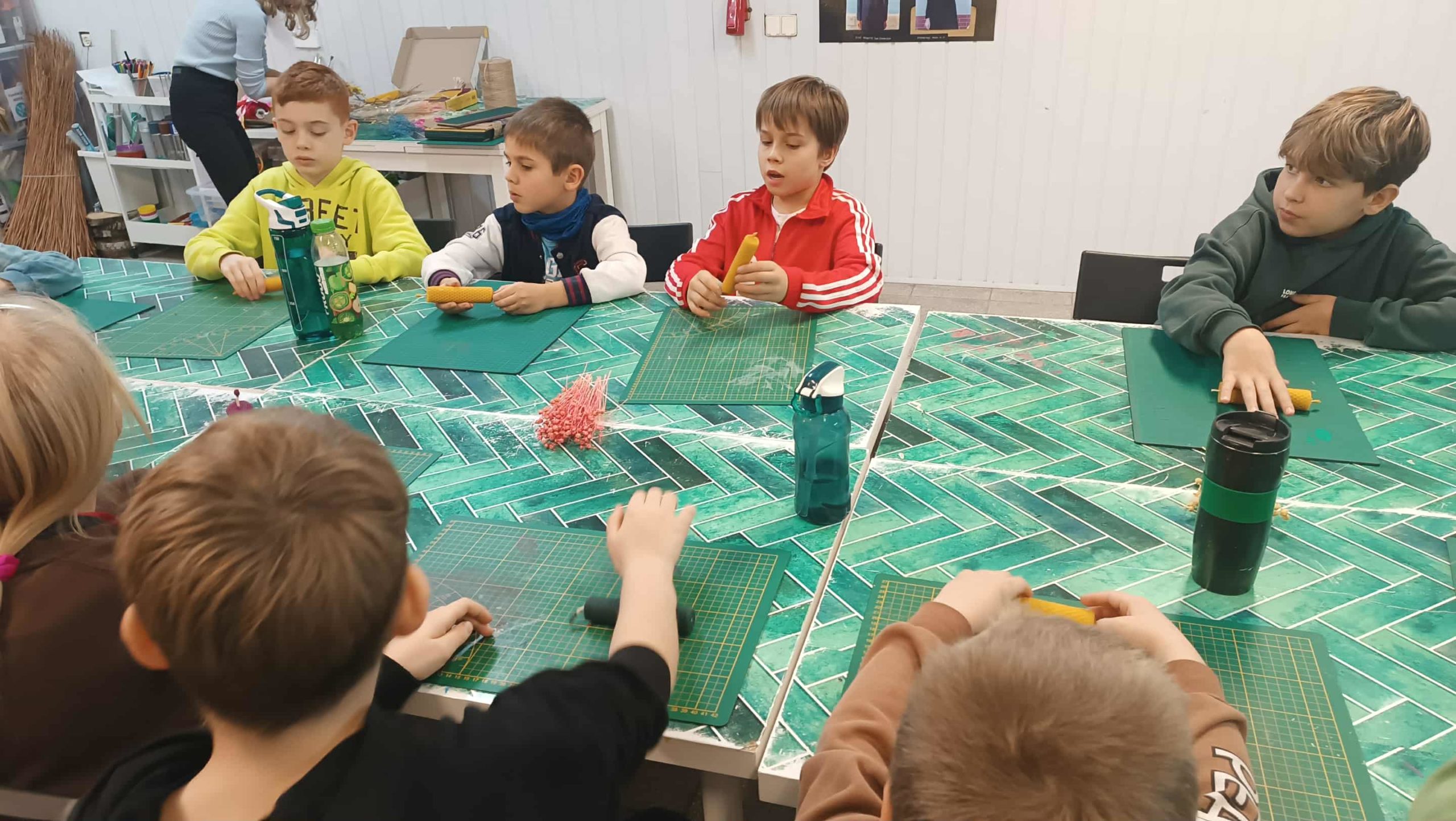 Grupa dzieci siedzi przy dużym stole pokrytym zieloną folią ochronną i pracuje nad rękodziełem. Każde dziecko ma przed sobą matę do cięcia i kawałek wosku ( węzy).  Na stole stoją też bidony i przybory plastyczne.