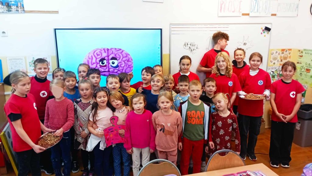 W sali lekcyjnej na tle tablicy multimedialnej z grafiką mózgu stoją ustawieni w trzy rzędy uczniowie klasy 1c i członkowie Koła PP HOPR. Pierwszy rząd uczniów trzyma plakat oraz tacę z orzechami