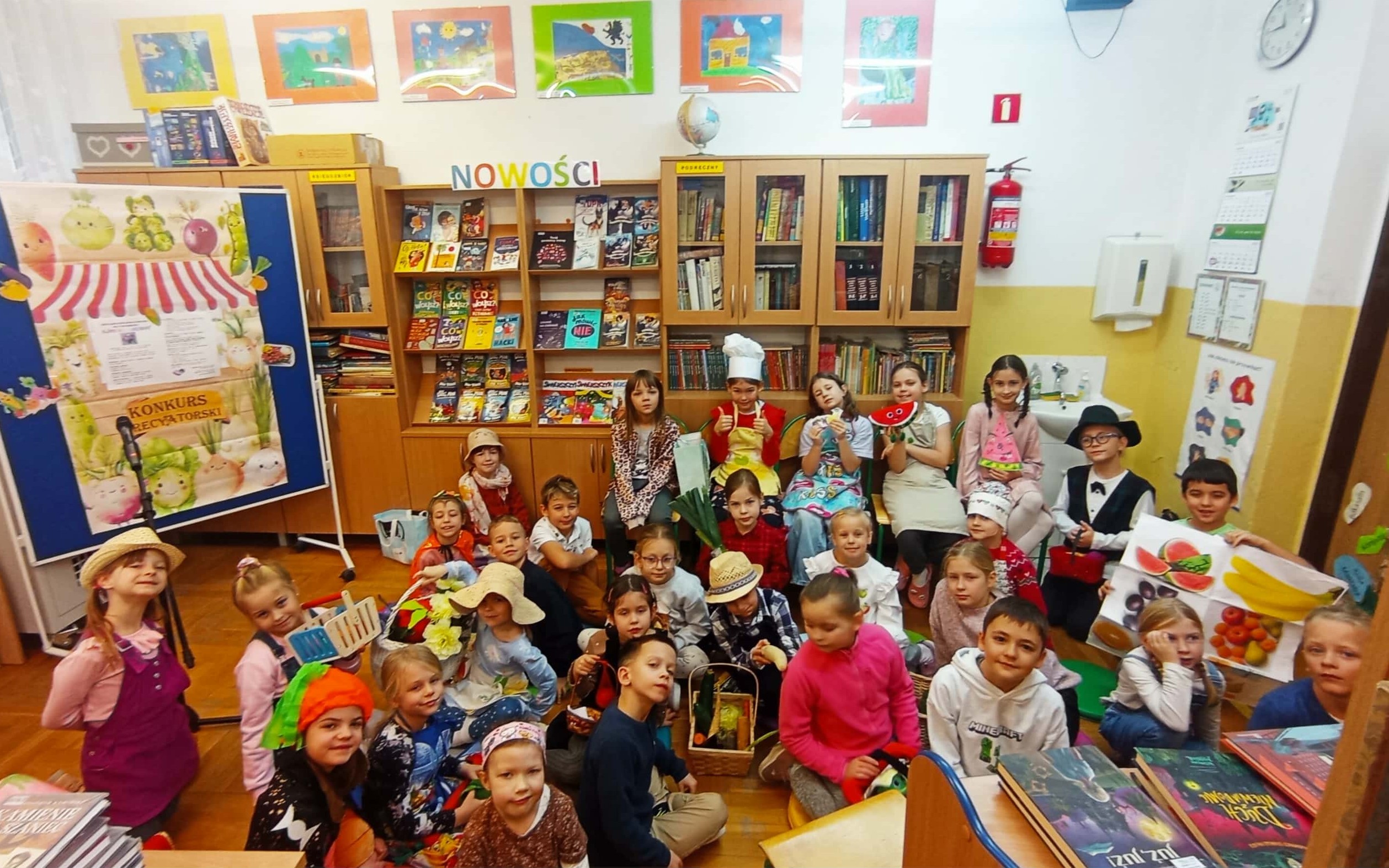 W bibliiotece szkolnej na tle dekoracji oraz regałów z książkami siedzi prawie 30 osobowa grupa uczniów klas I-III. Dzieci przebrane są w owocowo warzyne stroje, a w rękach trzymają rekwizyty.