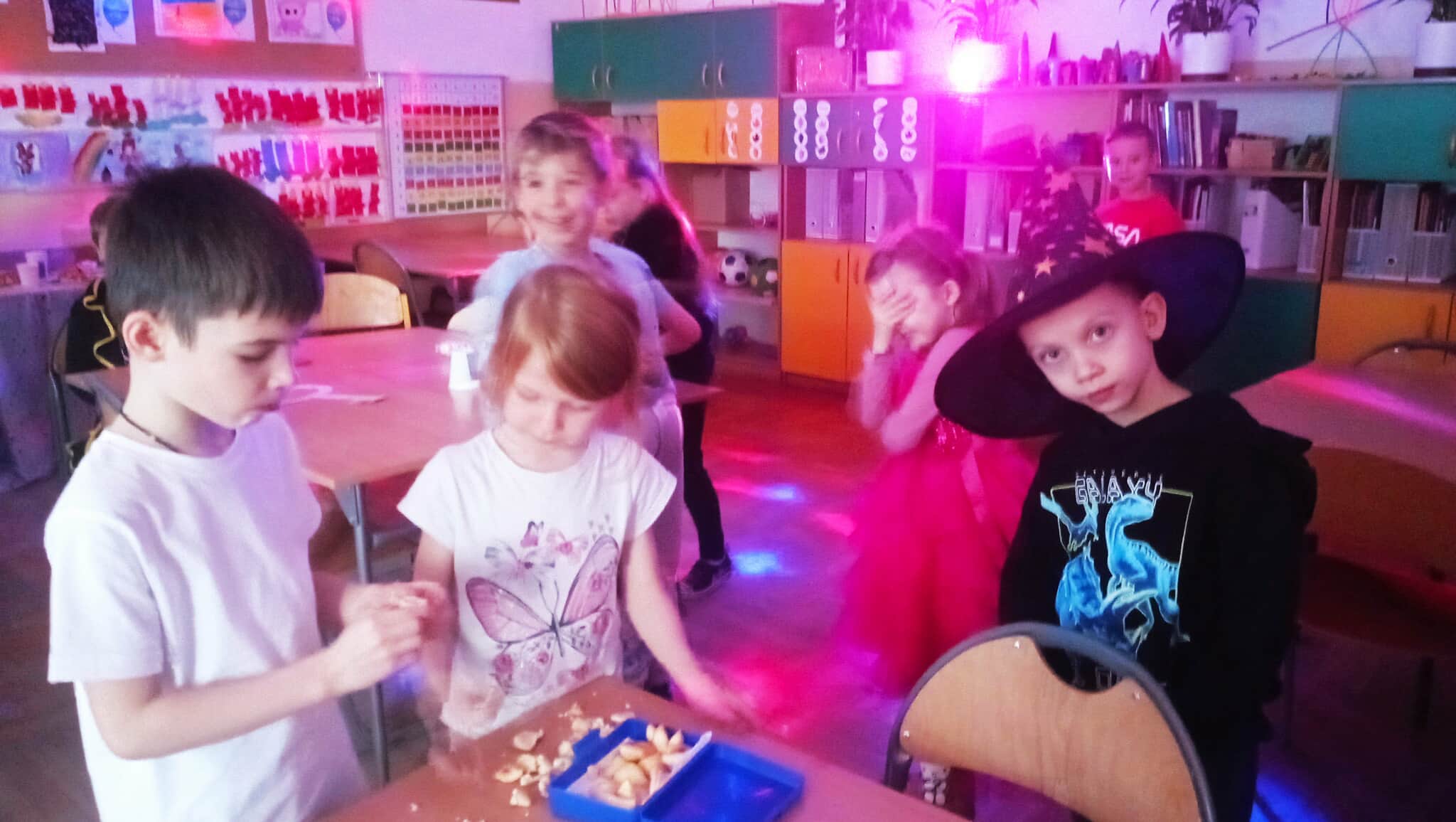 W sali lekcyjnej w koło stolika na tle kolorowej poświaty kuli disco na pierwszym planie dziewczynka z pudełkiem ciasteczek z wróżbami, chłopiec otwierający ciasteczko oraz chłopiec w kapeluszu wróża. W tle czwórka innych bawiących się dzieci