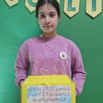 Na zdjęciu stoi dziewczynka w różowej bluzie, trzymająca żółte pudełko z napisem „Dzień Życzliwości – losy życzliwości do wylosowania”. W tle widać zieloną tablicę ozdobioną girlandą z literami.