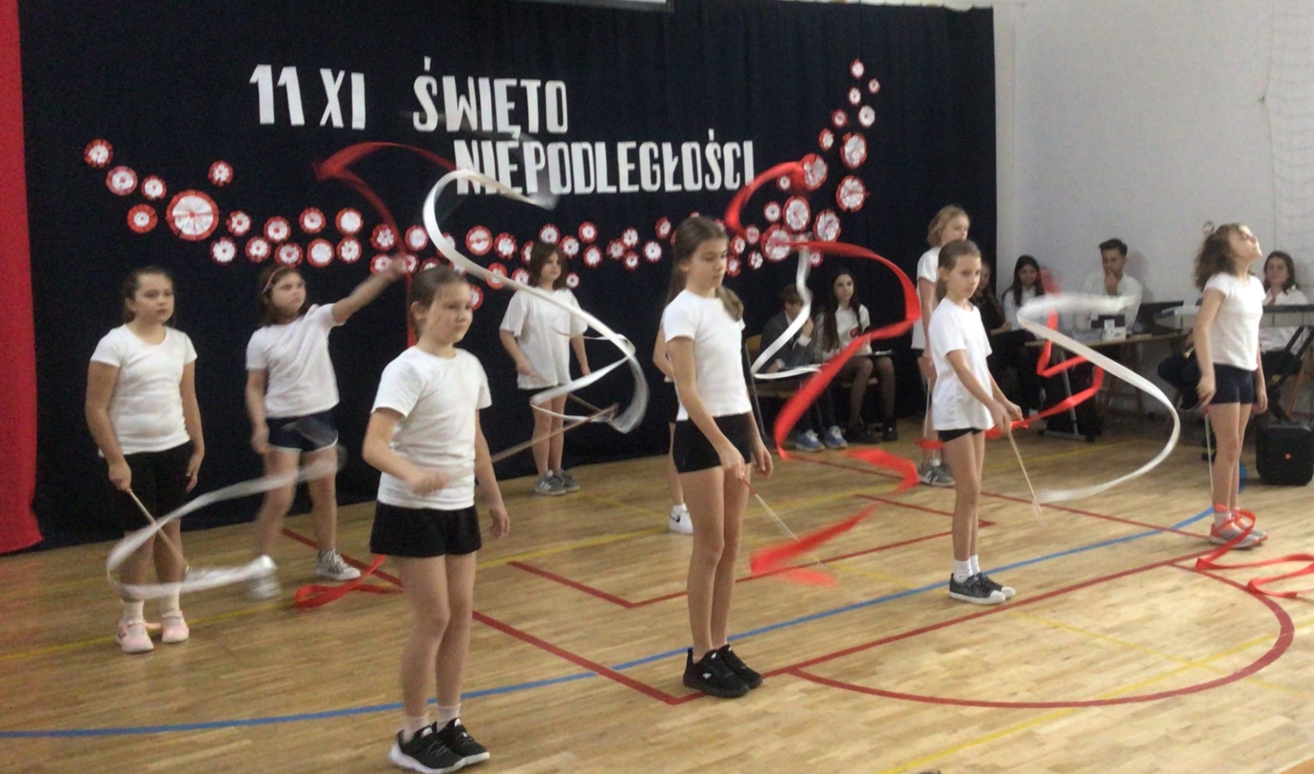 Na zdjęciu widać uczennice występujące w sali gimnastycznej podczas szkolnej akademii z okazji Narodowego Święta Niepodległości. Dziewczęta ubrane są w białe koszulki i czarne spodenki, a w rękach trzymają czerwone i białe szarfy, którymi wykonują układ taneczny. W tle znajduje się dekoracja z napisem „11 XI ŚWIĘTO NIEPODLEGŁOŚCI” oraz biało-czerwone elementy ozdobne.