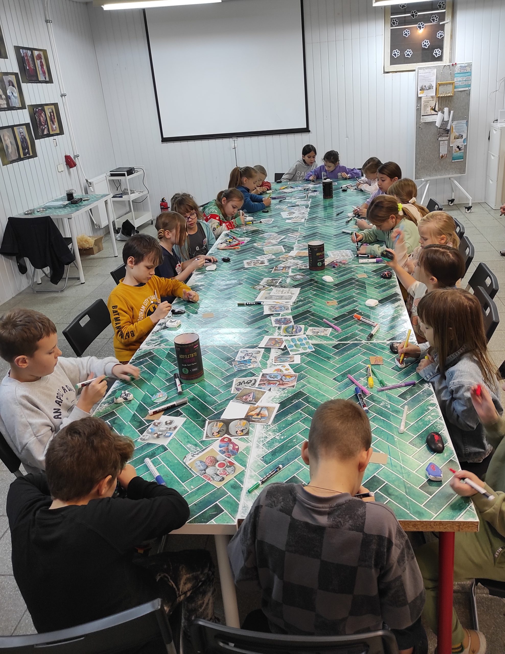 19 dzieci siedzi wokół stołu, który jest nakryty wzorzystą ceratą. Na stole leżą: okrągłe pojemniki, mazaki, obrazki.