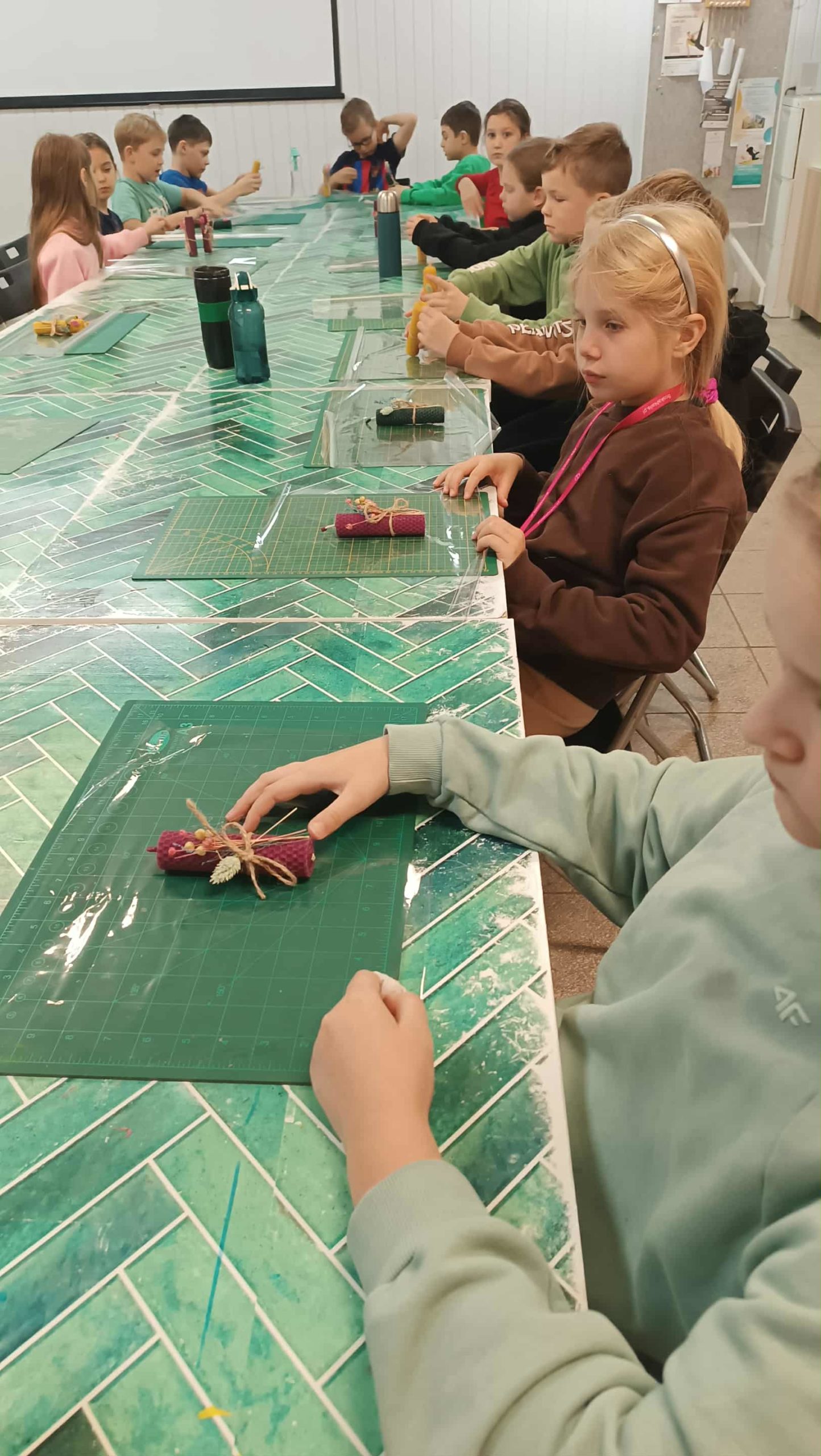 Dzieci siedzą przy stole, trzymając w rękach zrobione przez siebie świeczki z węzy pszczelej. Na matach leżą już gotowe, ozdobione świeczki przewiązane sznurkiem. Uczniowie wyglądają na zadowolonych z efektów swojej pracy.