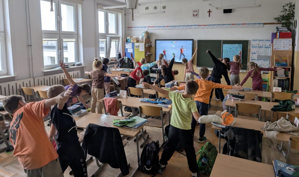 Uczniowie stoją obok swoich ławek i wykonują ćwiczenia ruchowe, naśladując postacie z filmu wyświetlanego na ekranie z przodu klasy