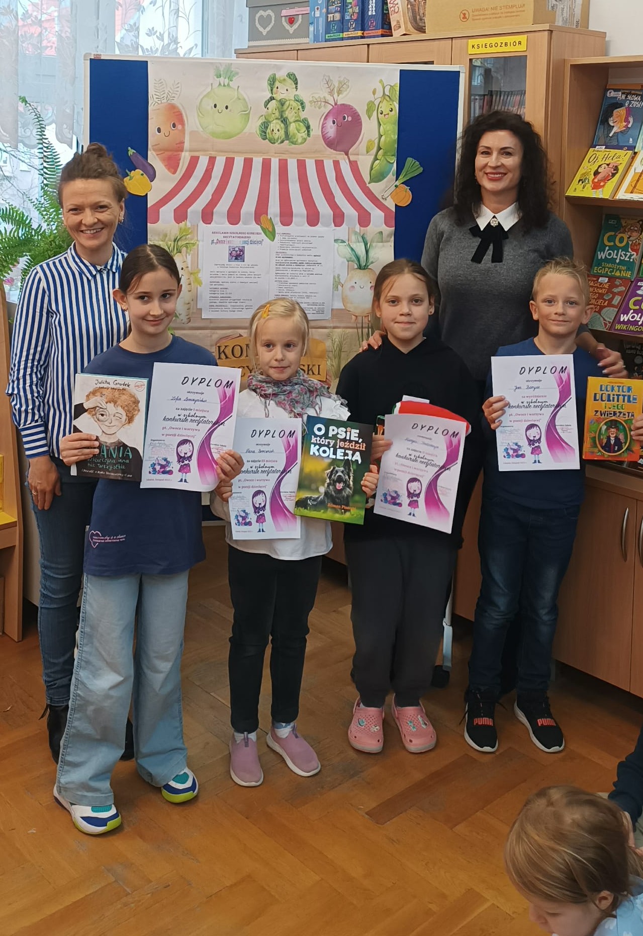 4 nagrodzonych uczniów wraz z dwiema nauczycielkami stoi na tle tablicy z dekoracją przedstawiającą warzywny stragan oraz z regulaminem konkursu recytatorskiego. W dłoniach trzymają nagrody książkowe wraz z dyplomami.