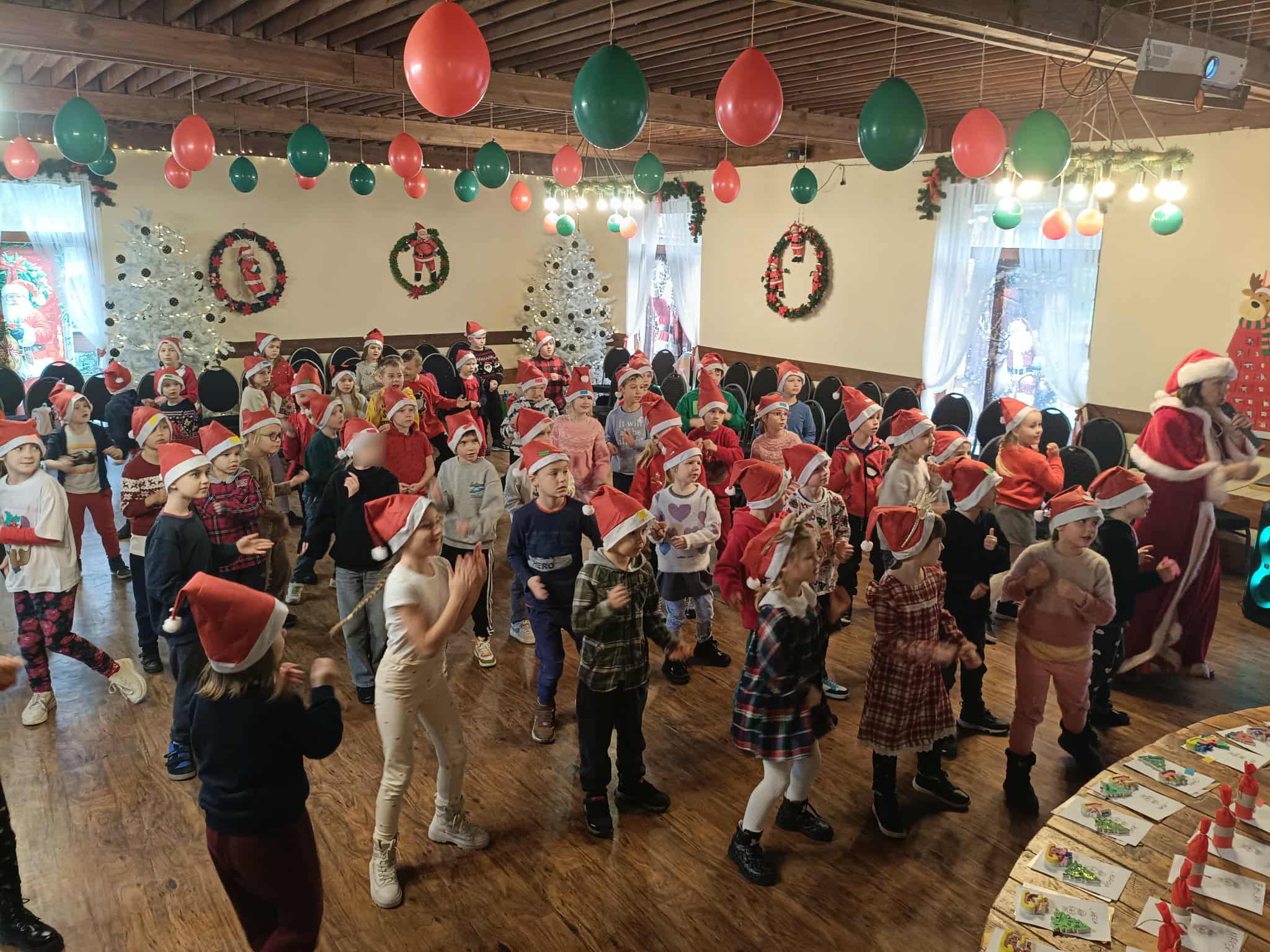 Duża grupa dzieci w czapkach Mikołaja bierze udział w zabawie ruchowej w udekorowanej świątecznie sali. Z sufitu wiszą czerwone i zielone balony, a w tle widać białe choinki i świąteczne wianki. Po prawej stronie stoi osoba w stroju świątecznym prowadząca zabawę.
