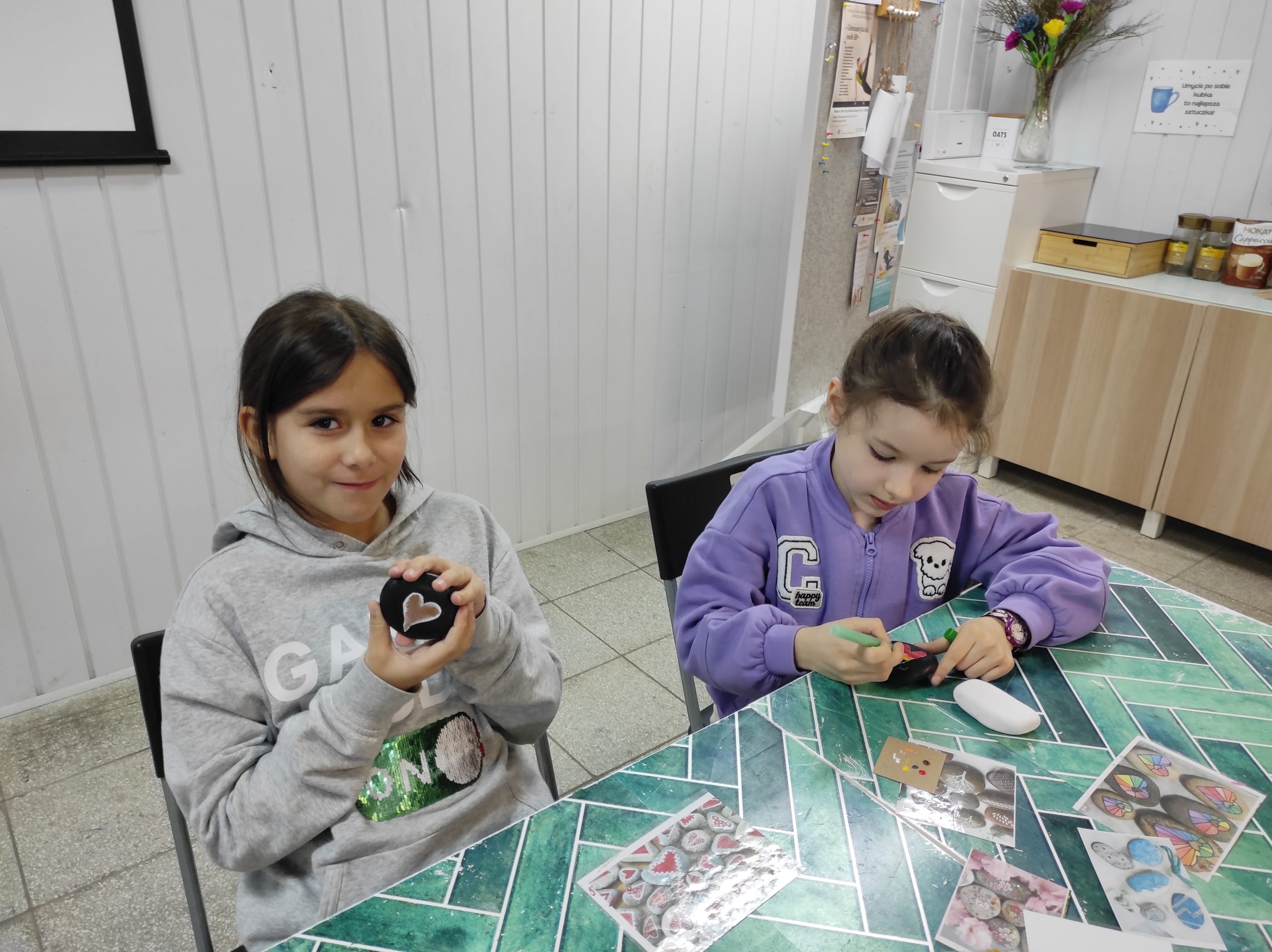 Na pierwszym planie dwie siedzące przy stole dziewczynki, jedna trzyma w dłoniach pomalowany kamień z serduszkiem, druga trzyma w prawej dłoni mazak, a lewą przytrzymuje czarny kamień. W tle elementy pomieszczenia.