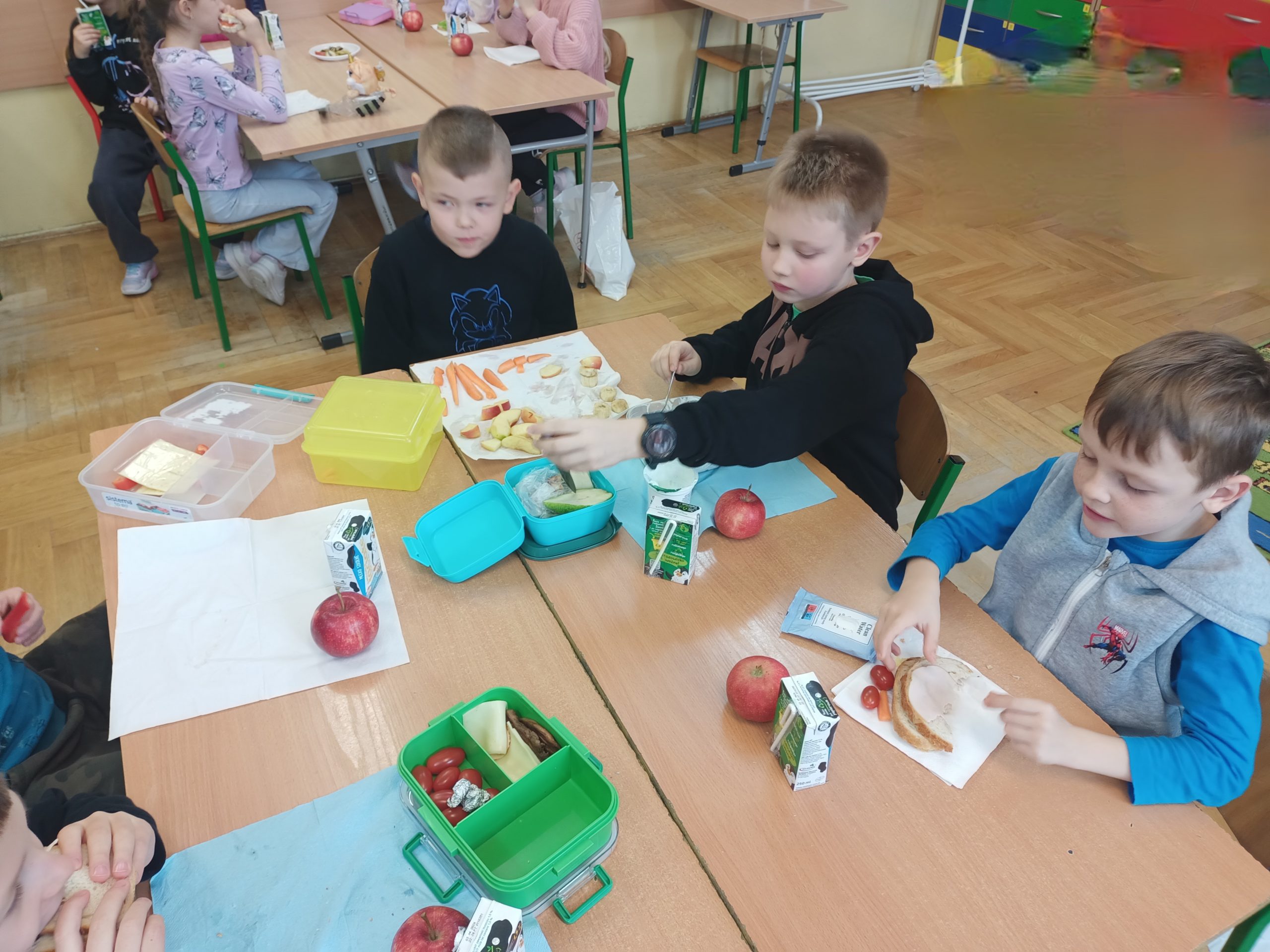 Troje uśmiechniętych uczniów siedzi w sali przy stolikach, przed nimi zdrowe produkty żywieniowe, bidony z wodą i śniadaniówki.