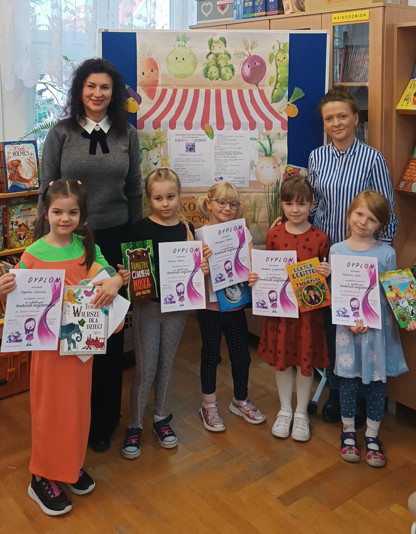 5 nagrodzonych uczennic wraz z dwiema nauczycielkami stoi na tle tablicy z dekoracją przedstawiającą warzywny stragan oraz z regulaminem konkursu recytatorskiego. W dłoniach trzymają nagrody książkowe wraz z dyplomami.