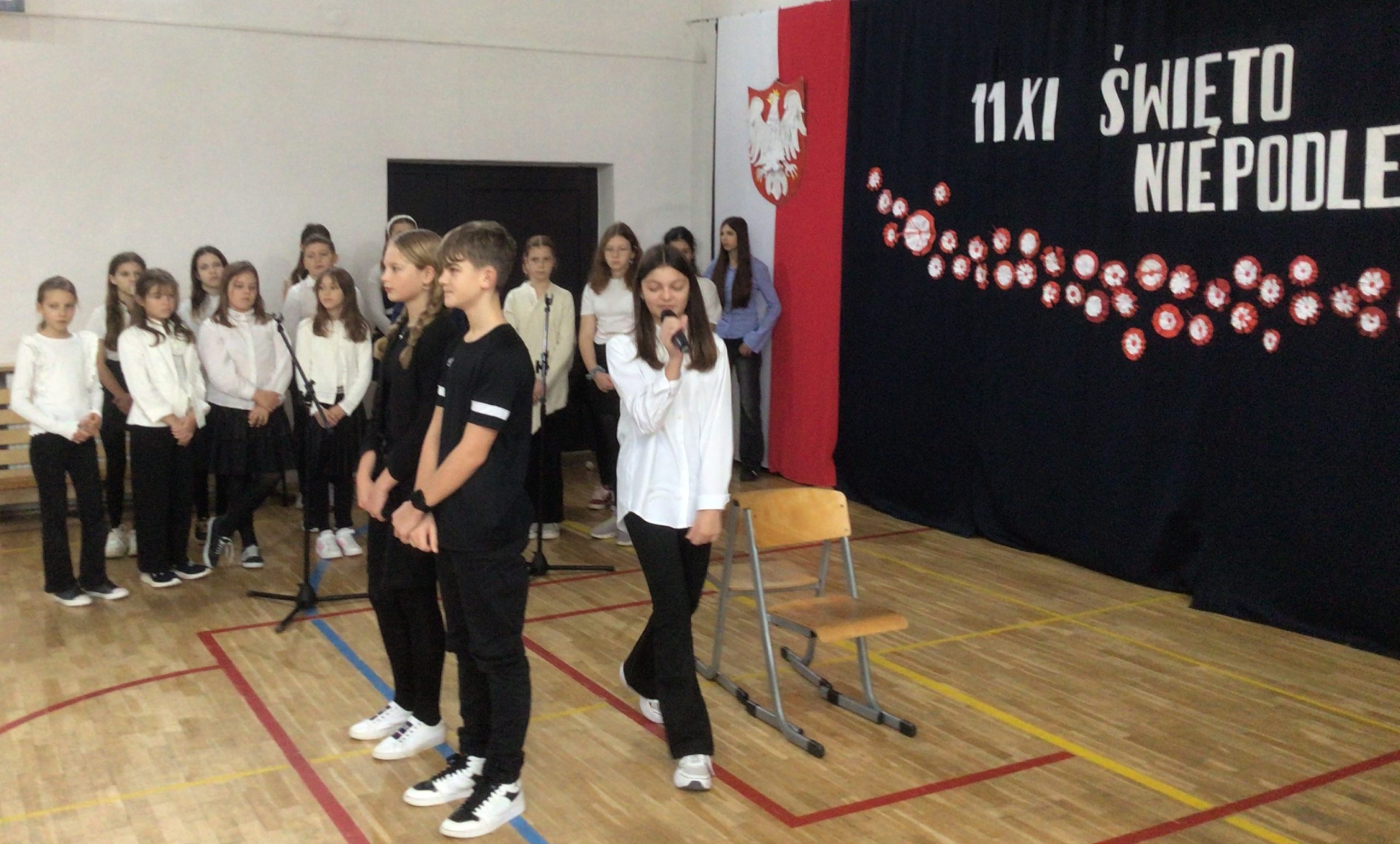 Na zdjęciu widać szkolną akademię z okazji Narodowego Święta Niepodległości obchodzonego 11 listopada. Na tle dekoracji w barwach narodowych – biało-czerwonej flagi i napisu „11 XI ŚWIĘTO NIEPODLEGŁOŚCI” – stoi grupa uczniów. Troje z nich znajduje się na pierwszym planie: dwie dziewczynki i chłopiec, z których jedna trzyma mikrofon i przemawia lub śpiewa. W tle stoją pozostali uczniowie, ubrani w odświętne stroje w kolorach czerni i bieli. Cała scena odbywa się w sali gimnastycznej szkoły.