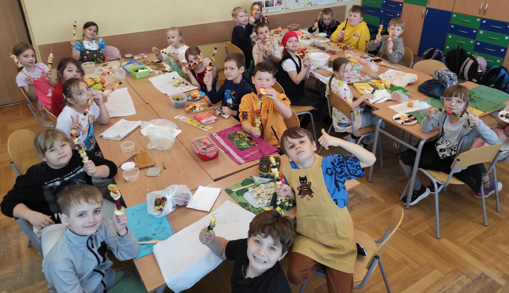 Grupa dzieci z 2 b siedzi  w klasie dookoła dwóch długich stolików.  Przed nimi leżą deski do krojenia i owoce z których robią szaszłyki. Dzieci są zadowolone. W dłoniach trzymają gotowe szaszłyki.