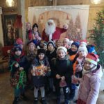  Grupa uśmiechniętych dzieci pozuje do zdjęcia z Mikołajem i elfem i nauczycielką w świątecznie udekorowanym, drewnianym wnętrzu. Dzieci trzymają małe prezenty, niektóre mają zimowe czapki i kurtki, a w tle widać choinkę oraz dekoracje świąteczne.