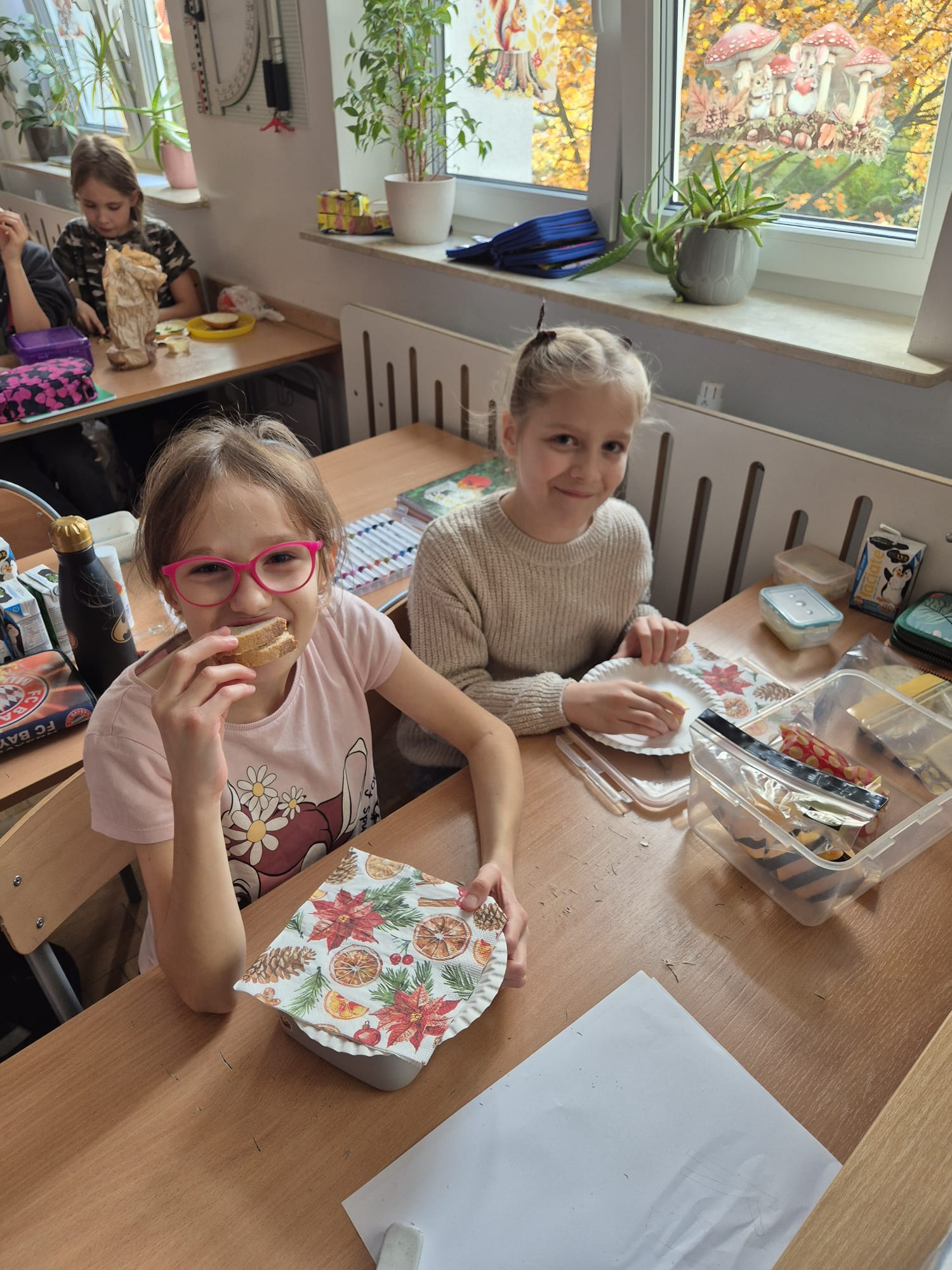 Dwie dziewczynki siedzą przy stole na który  są rozłożone produkty żywnościowe. W tle przechodzi chłopiec.