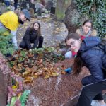 Grupa młodych osób porządkuje grób na cmentarzu jesienią. Na nagrobku widać liście i rośliny, a wokół leżą opadłe liście.