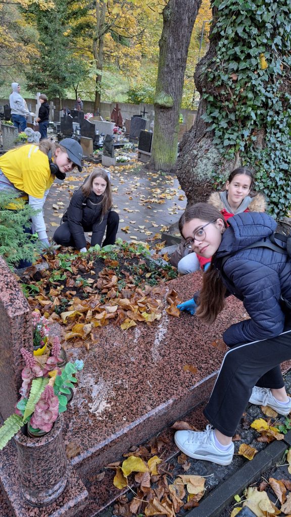 Grupa młodych osób porządkuje grób na cmentarzu jesienią. Na nagrobku widać liście i rośliny, a wokół leżą opadłe liście.