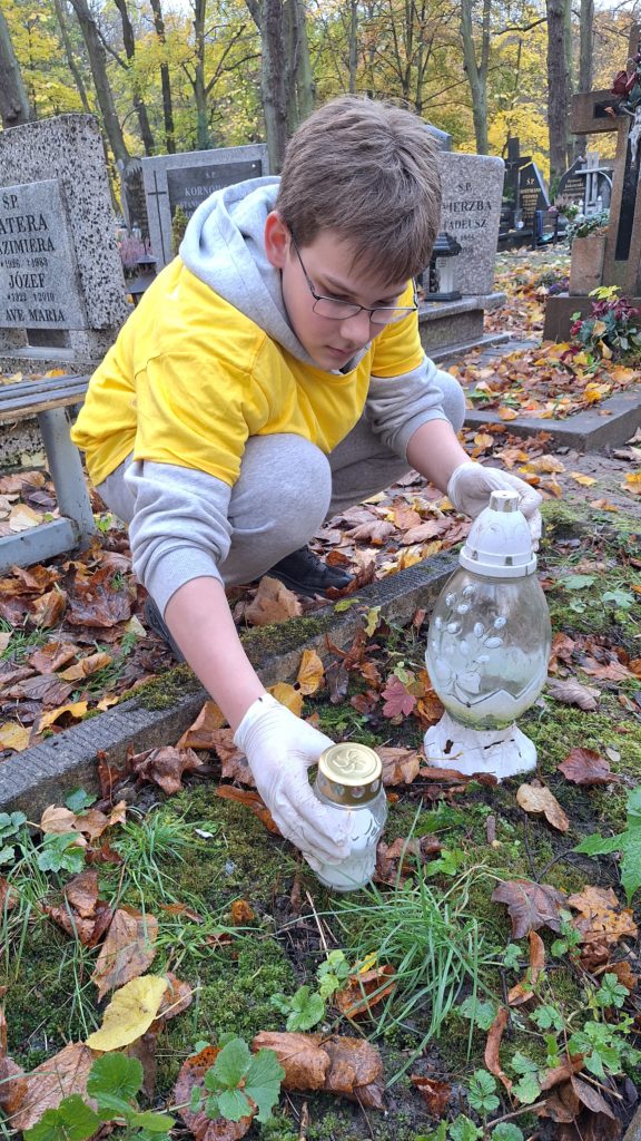 Dziewczynka w koszulce wolontariusza i czapce sprząta grób na cmentarzu. Obok stoją znicze, a ziemię pokrywają jesienne liście.
