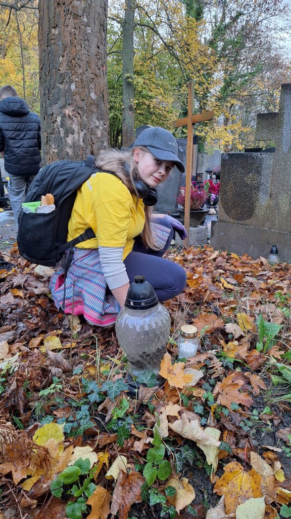 Dziewczyna w koszulce wolontariusza i czapce z daszkiem kuca przy grobie, na którym leżą jesienne liście. W tle stoi drewniany krzyż, przed dziewczyną duży znicz.