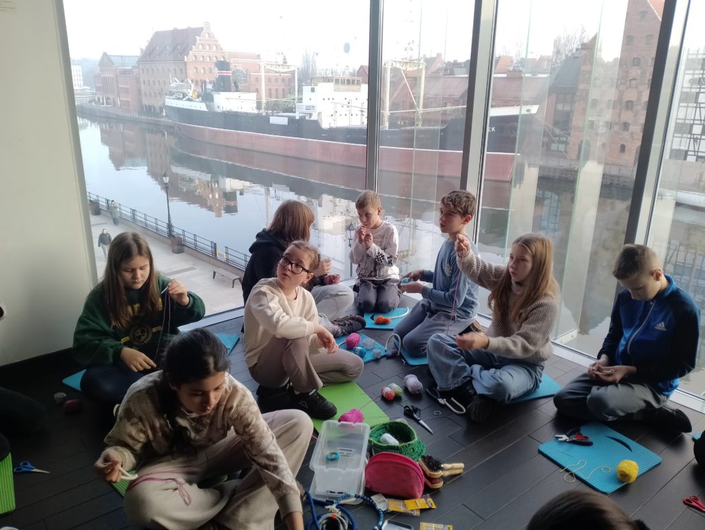 Grupa dzieci siedzi na podłodze przy dużych oknach z widokiem na Motławę, zabytkowe budynki i statek. Dzieci zajmują się pracą manualną, trzymają włóczki, nożyczki, sznurki.