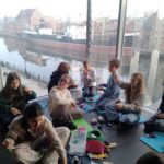 Grupa dzieci siedzi na podłodze przy dużych oknach z widokiem na Motławę, zabytkowe budynki i statek. Dzieci zajmują się pracą manualną, trzymają włóczki, nożyczki, sznurki.