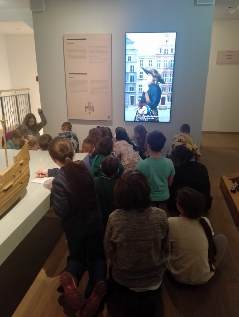 Grupa dzieci siedzi na podłodze i wykonuje zadania na kartkach. Obok dzieci znajduje się fragment dużego modelu statku. Na ścianie jest plansza informacyjna i ekran z postacią w historycznym stroju.