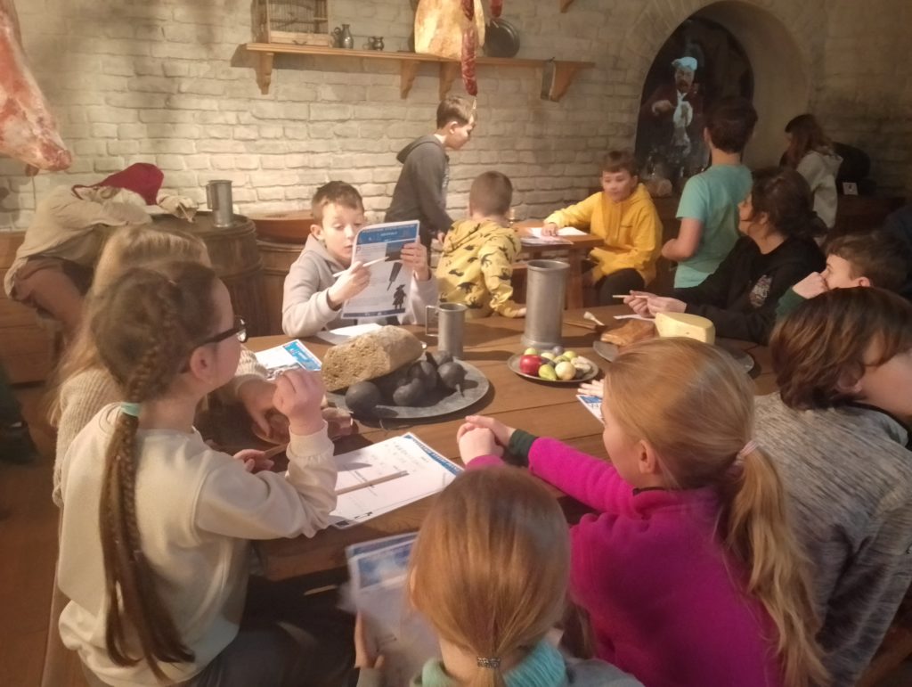 Grupa dzieci siedzi przy dużym drewnianym stole w pomieszczeniu muzealnym, stylizowanym na dawną kuchnię. Na stole leżą chleb, ser, owoce oraz metalowe naczynia. Dzieci trzymają w ręku kartki, ołówki. W tle stoją inne dzieci.