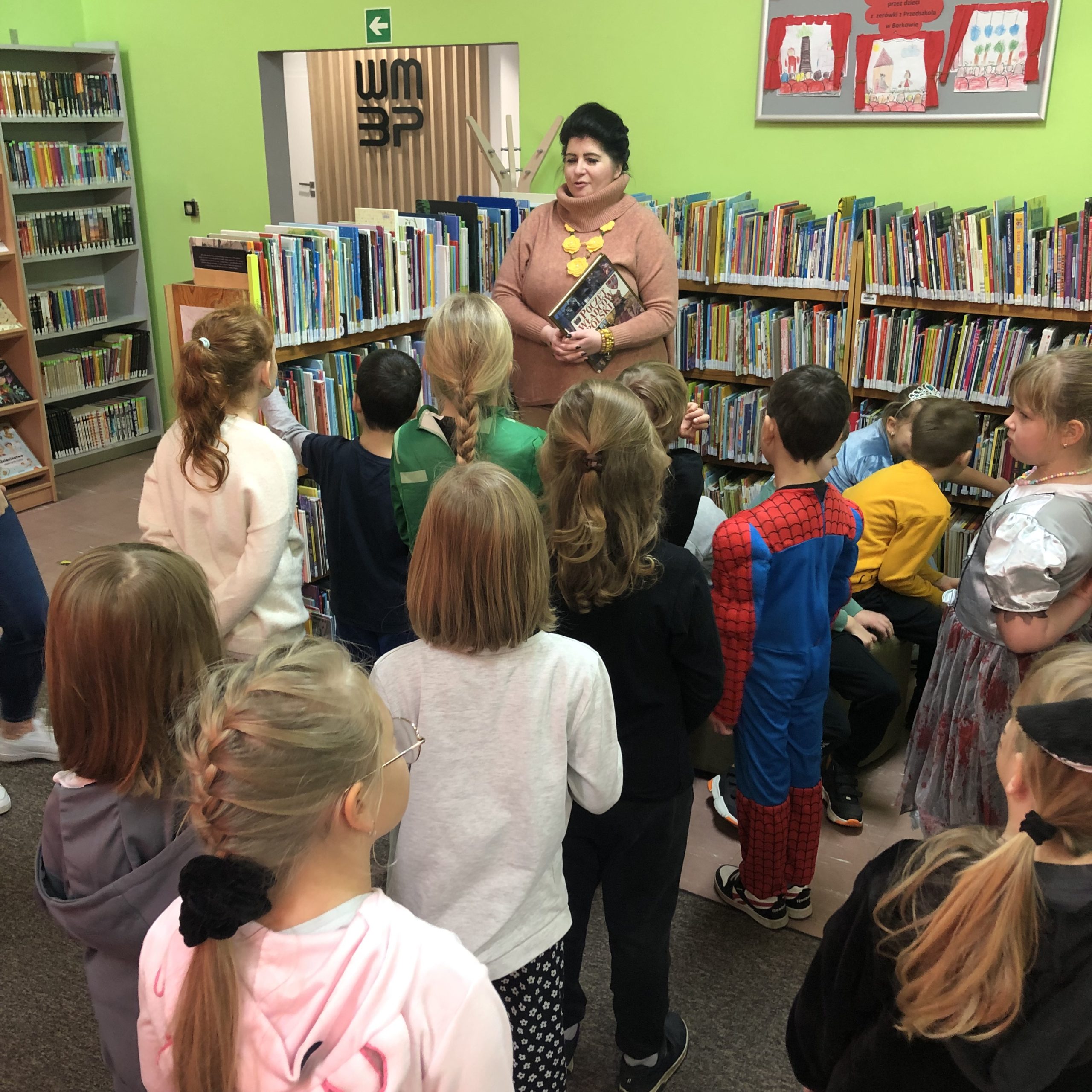 Grupa uczniów stoi zwrócona twarzą w stronę pani bibliotekarki. W tle są regały z książkami.