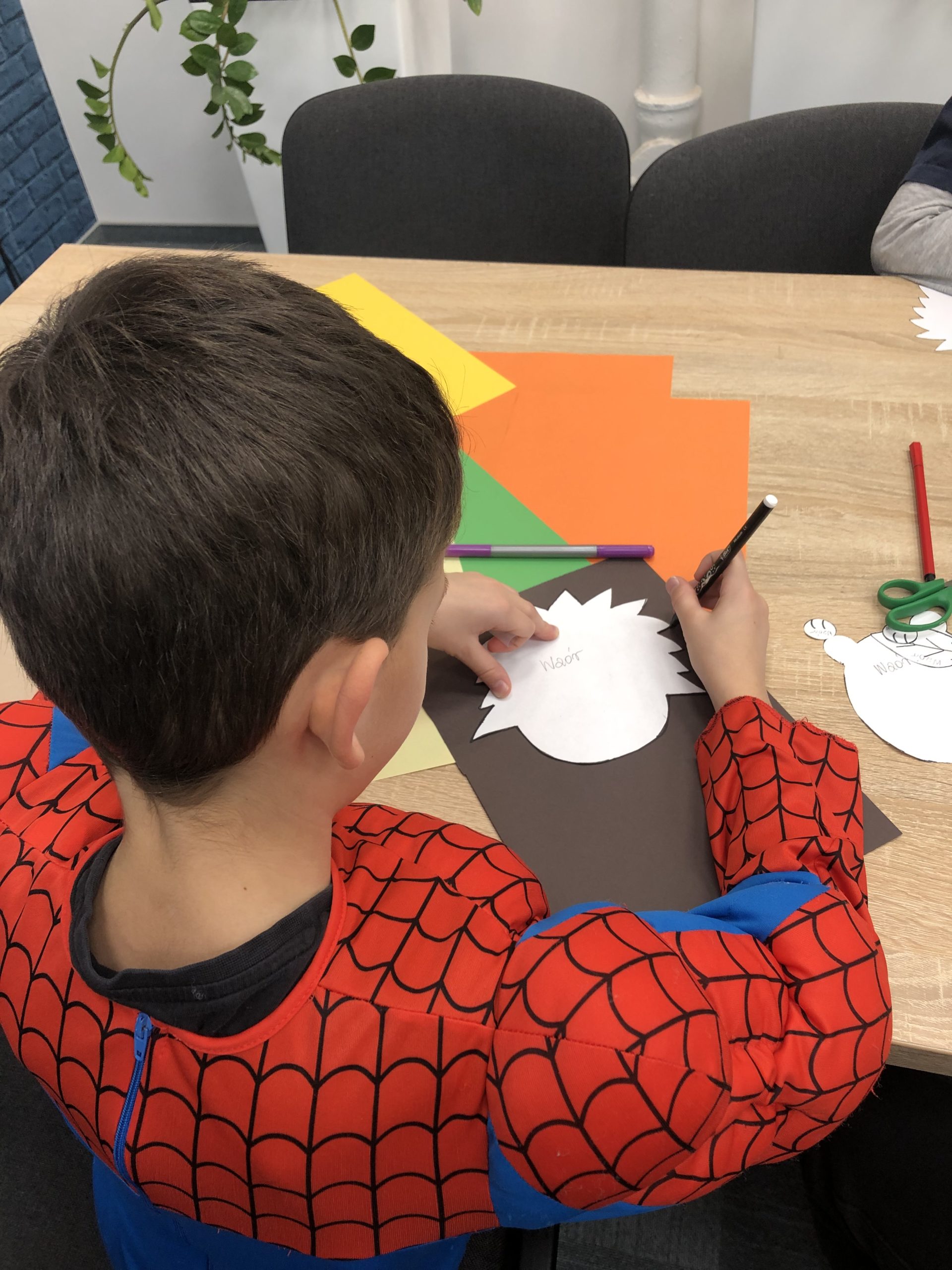 Chłopiec siedzi przy stole, przed nim kolorowe kartki i szablon jeża.